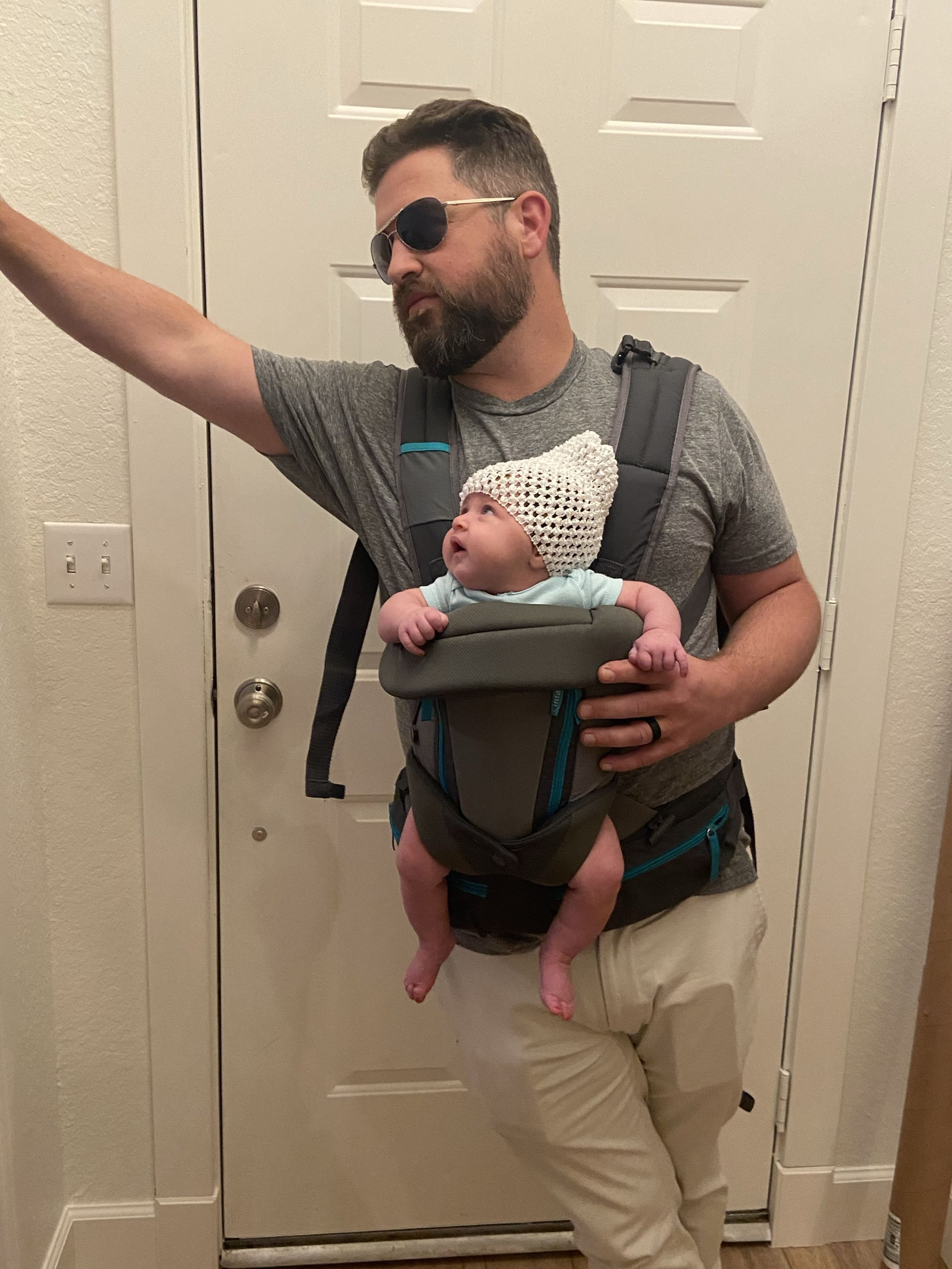 A man with a beard and sunglasses wearing a gray t-shirt, beige pants, and a backpack holding a baby in a front carrier in front of a white door. The man is pointing or gesturing with his left arm.