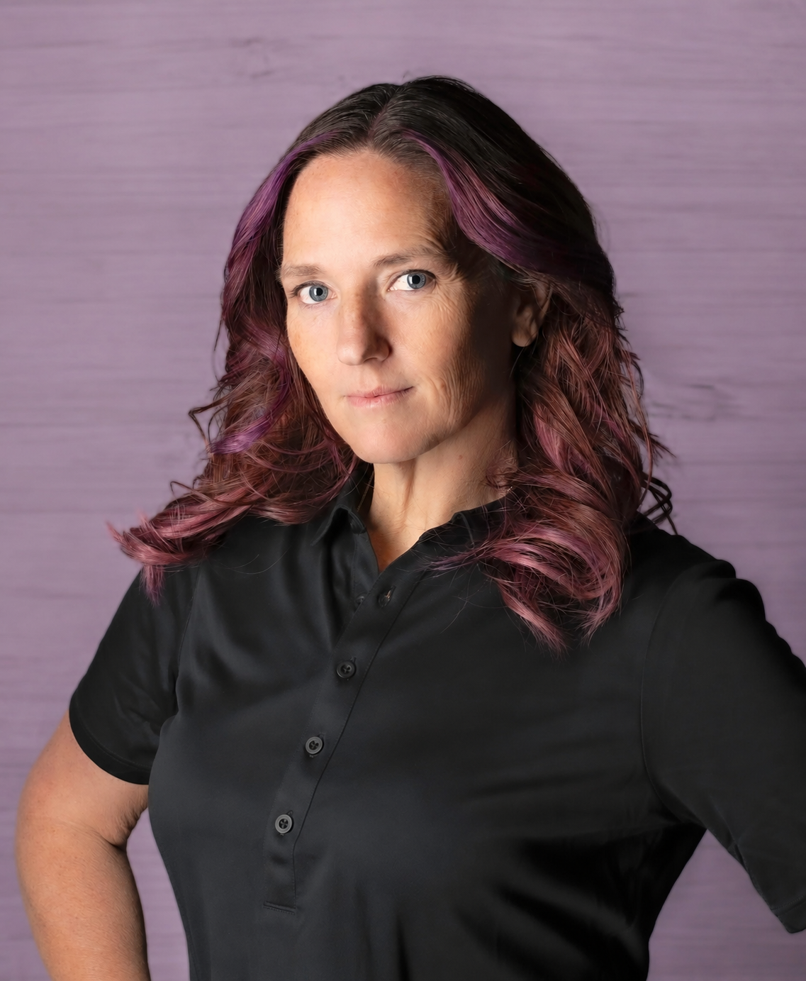 Woman with purple and black wavy hair wearing a black button-up shirt, standing against a purple background.