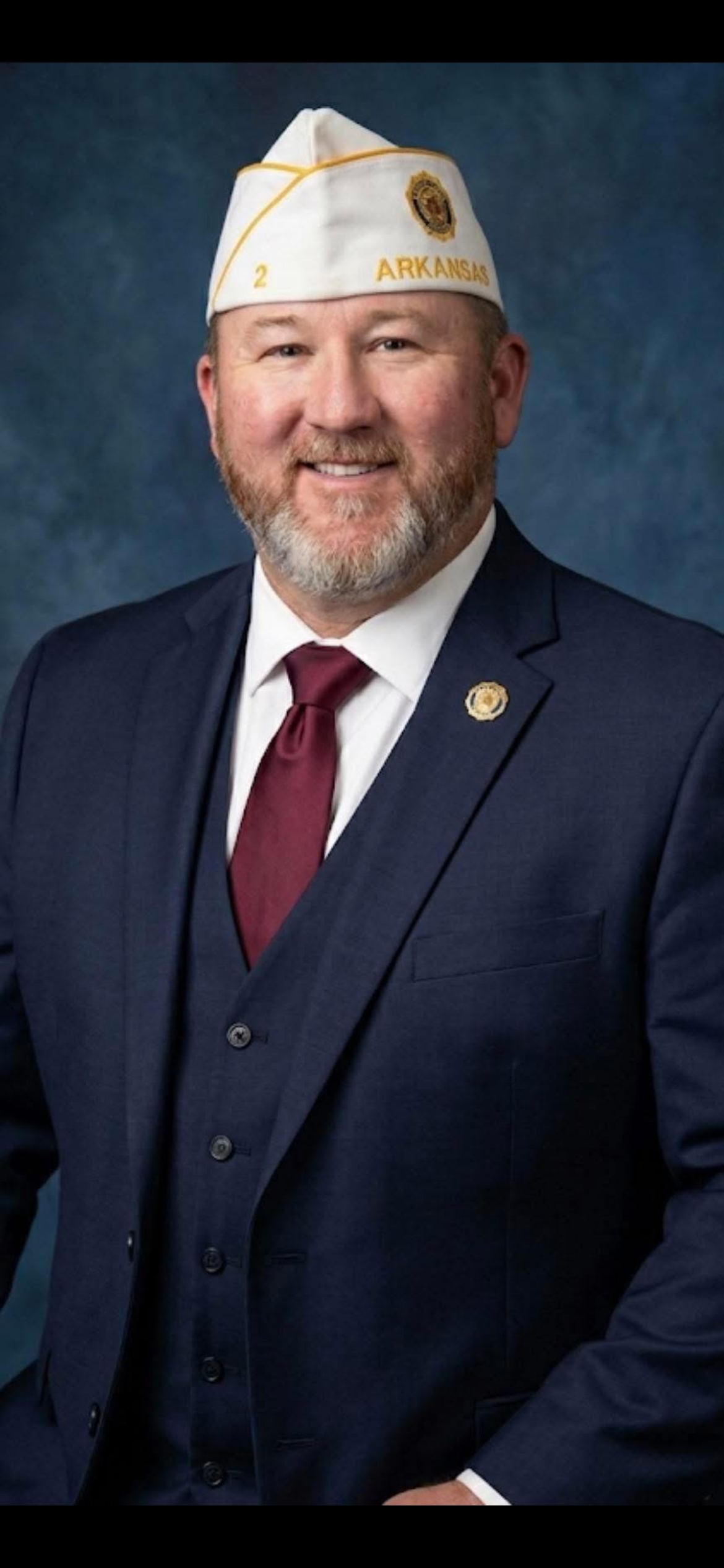 A man in a navy blue suit with a red tie and white shirt, wearing a white American Legion cap with gold trim and the words 'Arkansas' and a badge emblem, posing for a professional portrait against a blue background.