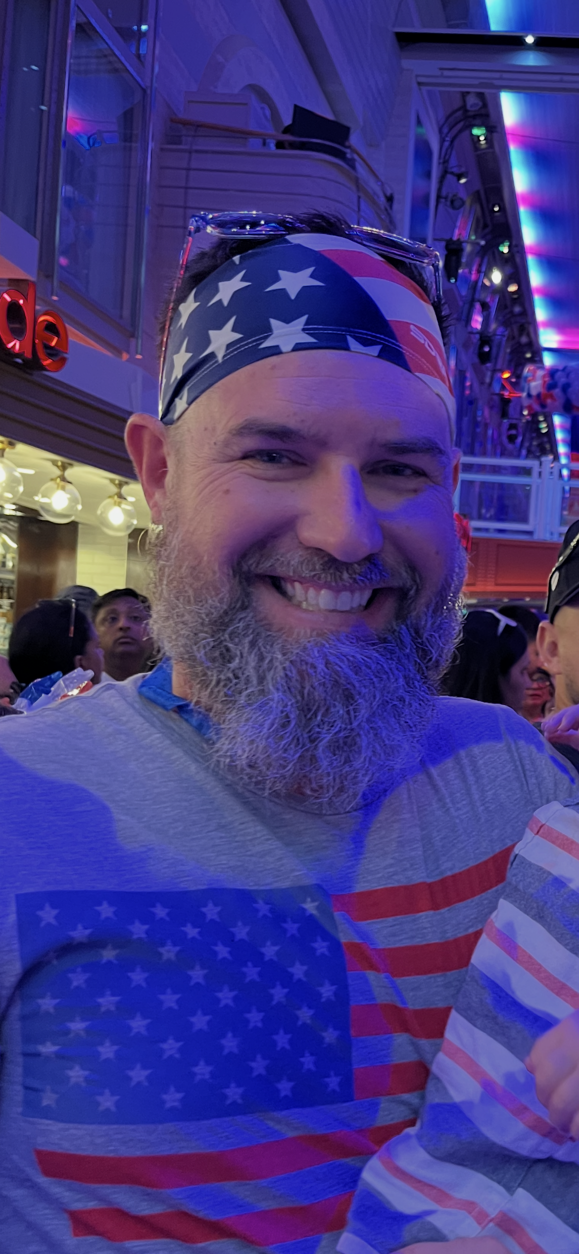 A smiling man with a beard wearing a bandana with American flag stars and stripes at a lively indoor event with colorful lighting.