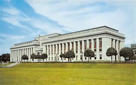 Large government building with columns, trees in front, and a grassy lawn under a partly cloudy sky.