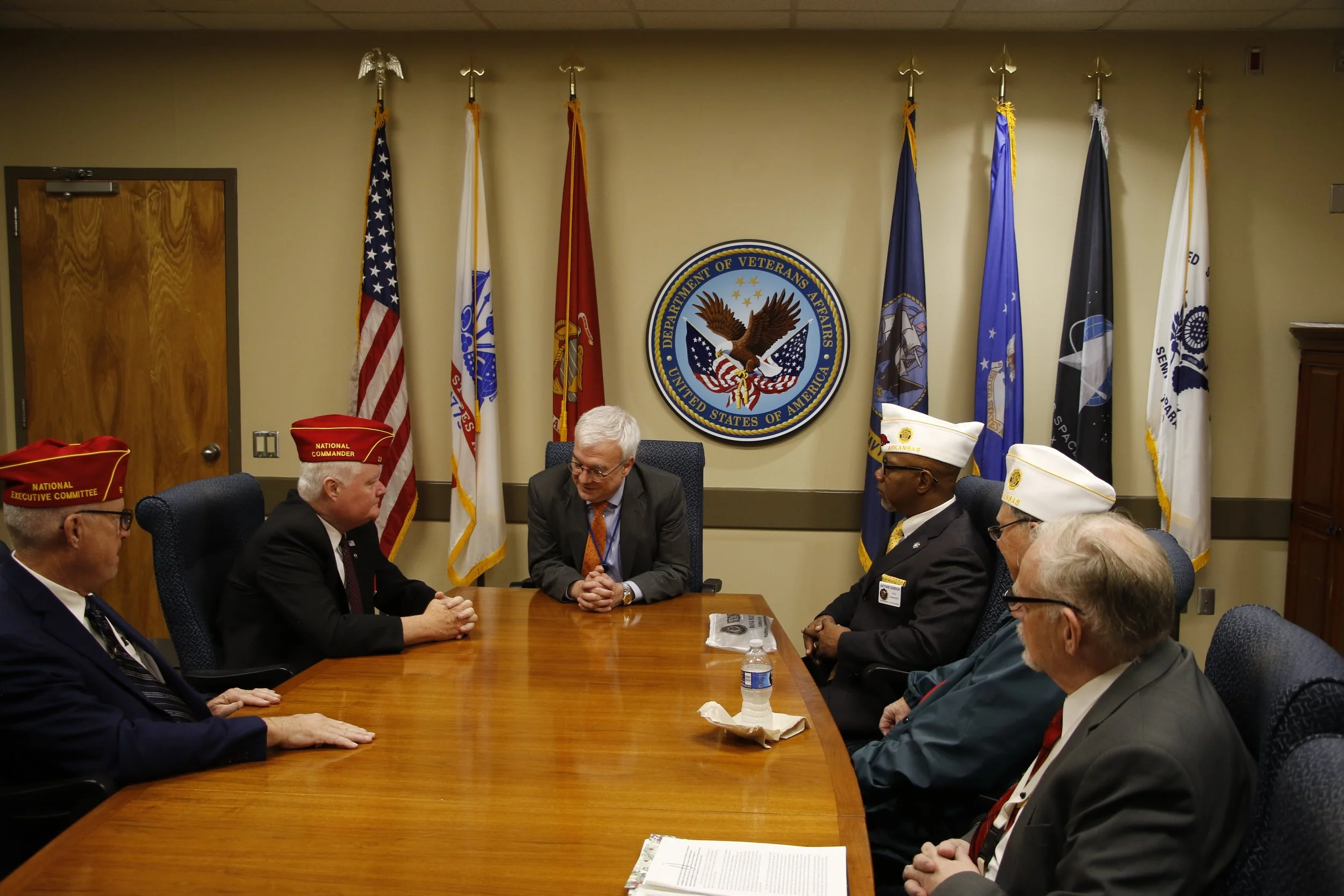Veterans and officials sitting around a conference table in a room with flags and a Department of Veterans Affairs emblem on the wall.