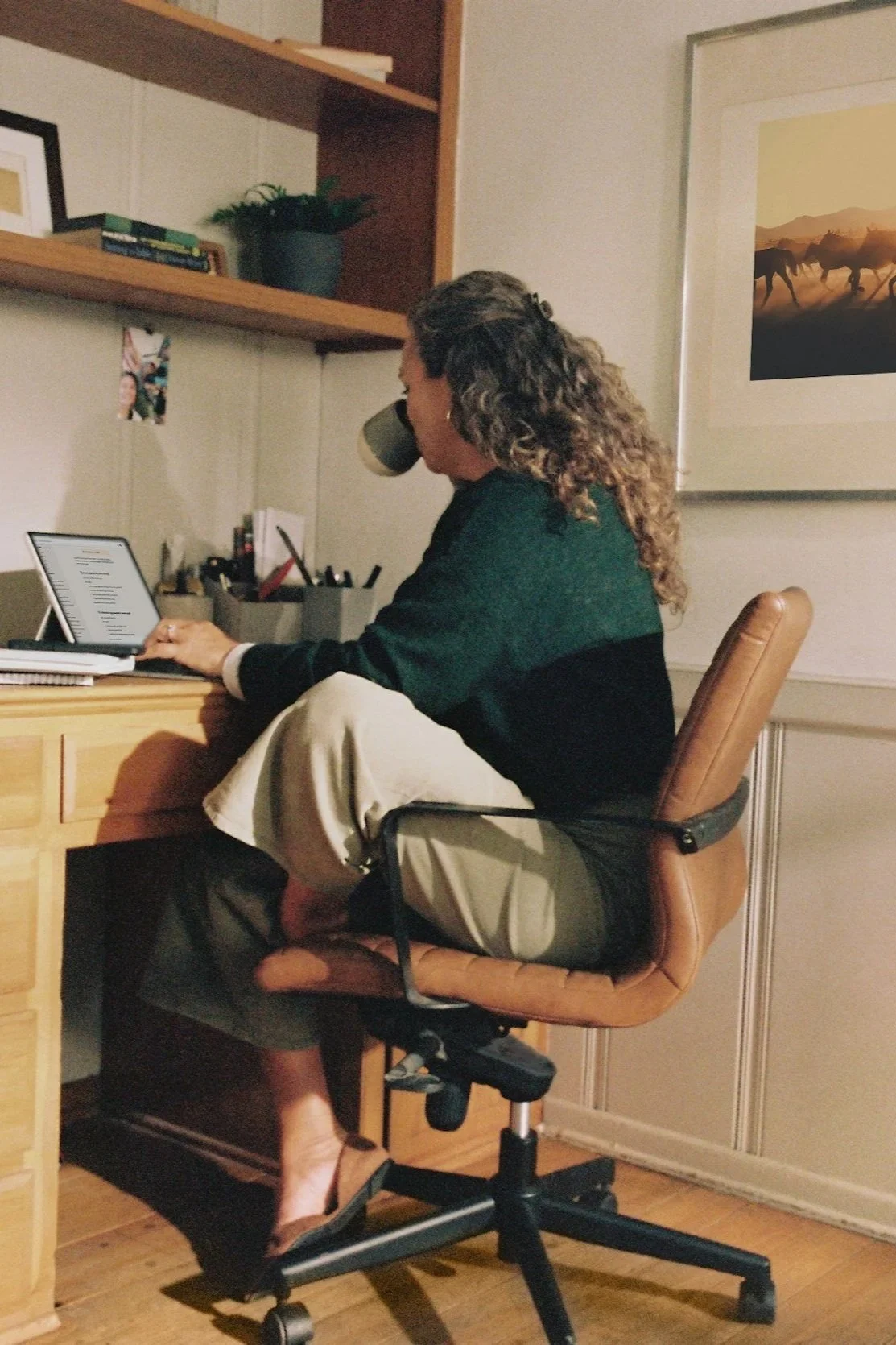 A woman sitting at a wooden desk, drinking from a mug while using a tablet. She is in an office or study room with shelves, framed art, and a painting of horses in the background.