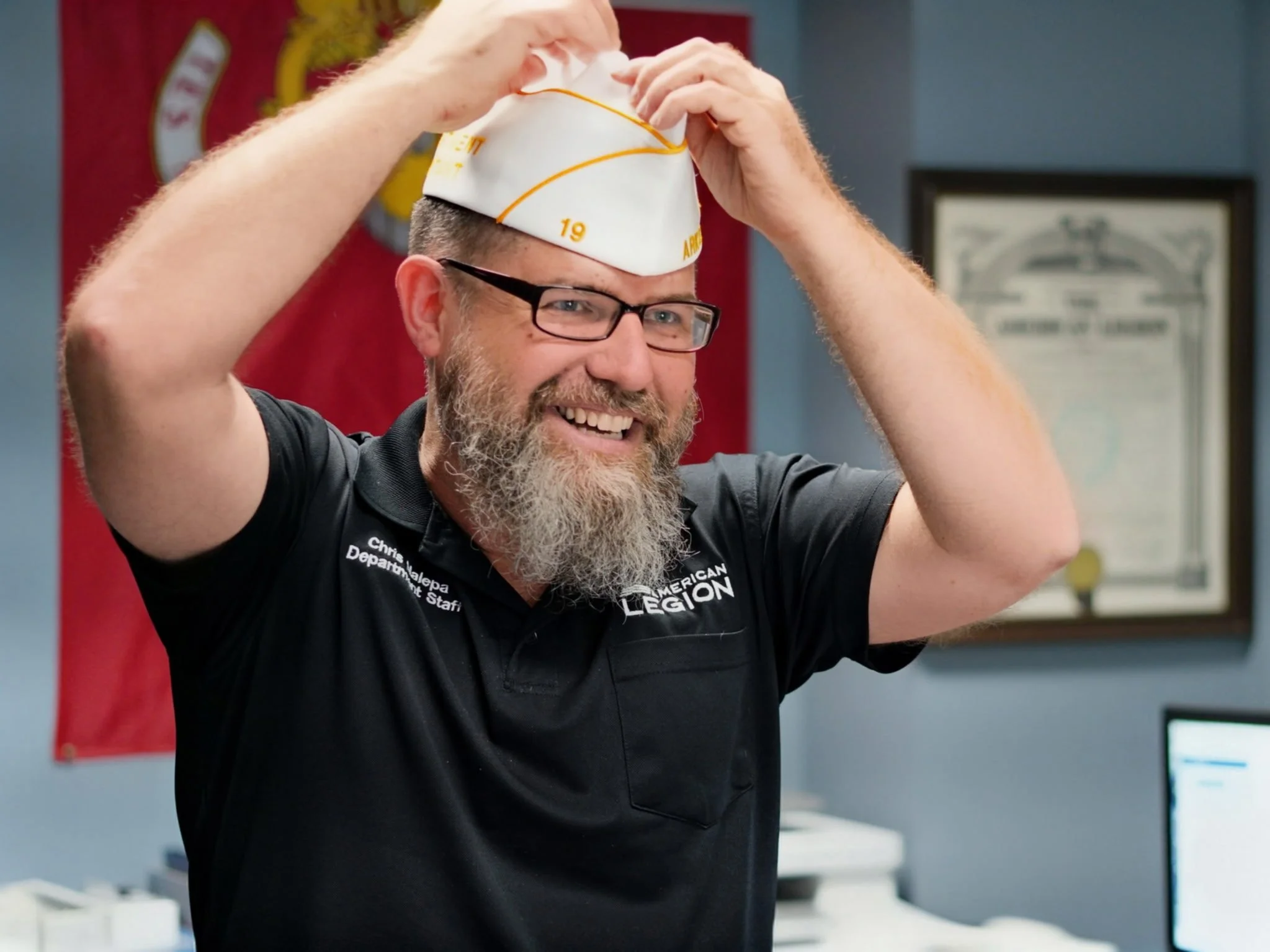 A smiling man with glasses and a beard putting on a white cap. He is standing indoors, possibly in an office or a meeting room, wearing a black polo shirt with white text.