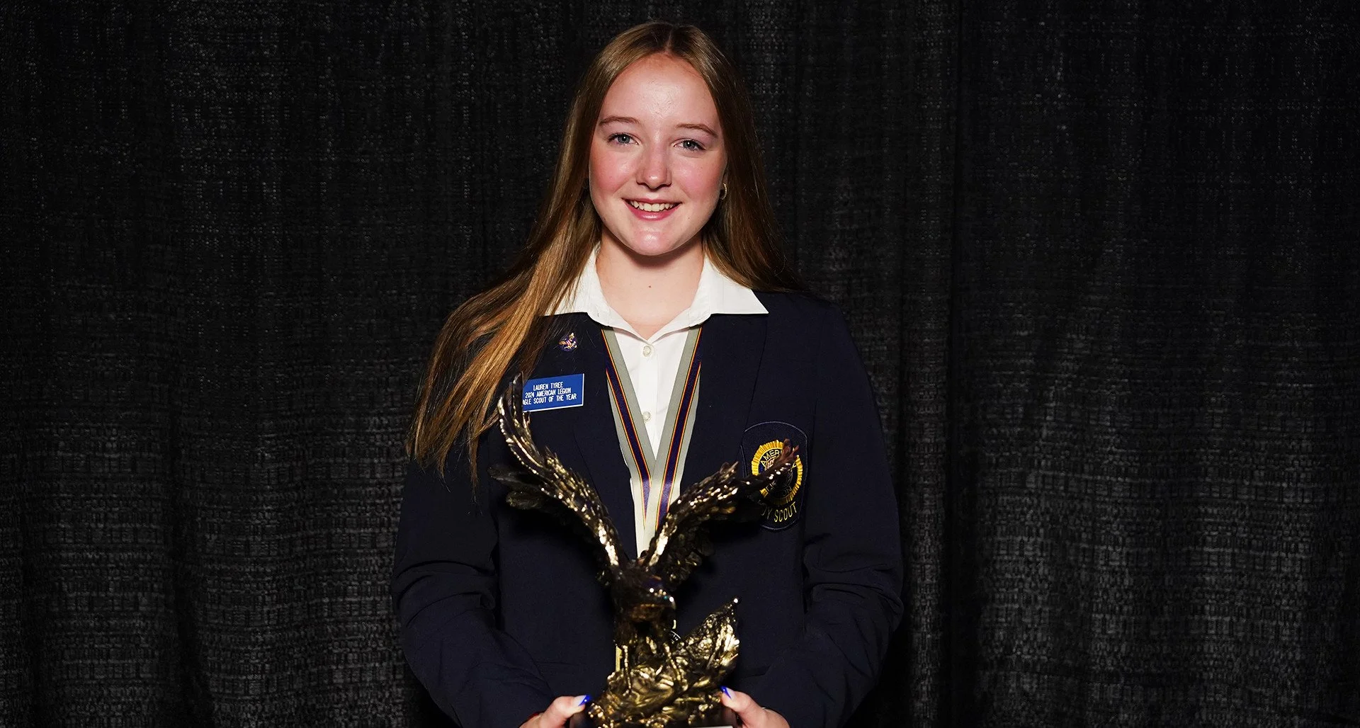 Young woman in Scout uniform holding an eagle-shaped award, standing against a black curtain background.