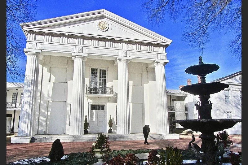 White neoclassical building with tall columns, a triangular pediment, and a fountain in the foreground, under a clear blue sky.