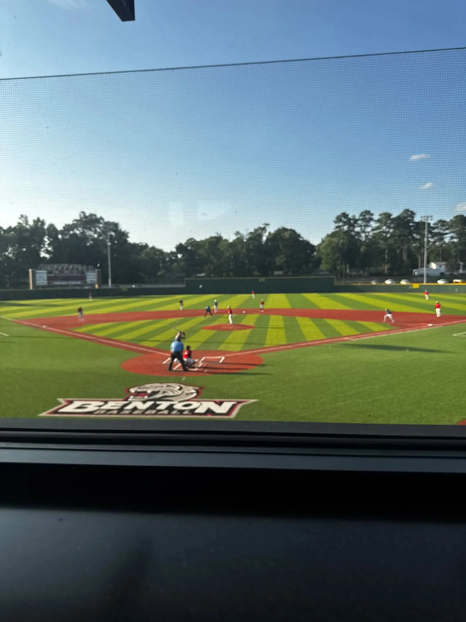 View of a baseball field during a game inside a stadium, seen through a window screen. The field has players, and there is an electronic scoreboard and trees in the background. A large logo with the words 'Envy of the Nation' is on the field.