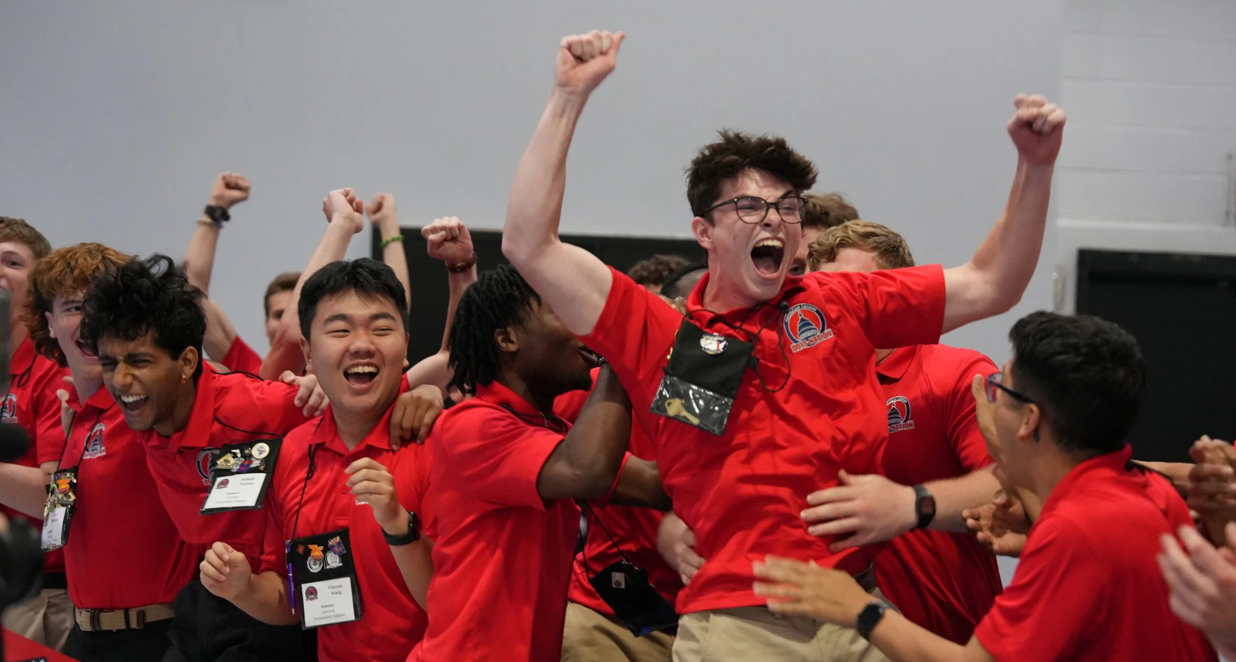 A group of young people in red shirts celebrating with excitement and joy, some with arms raised and smiling widely, indoors at a conference or event.