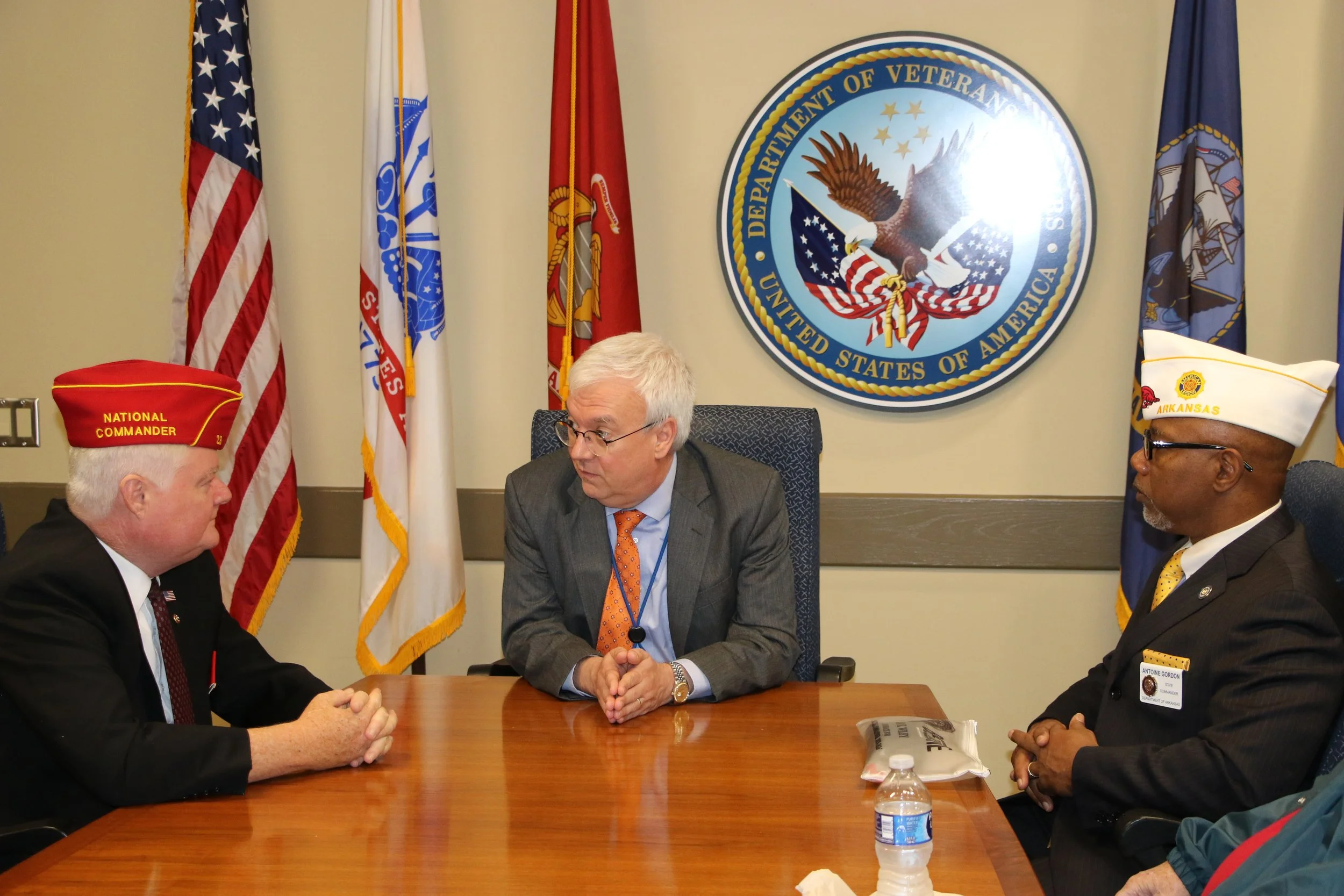Four men in formal attire sit around a conference table in a room decorated with flags and a large Department of Veterans Affairs emblem on the wall. The men are engaged in conversation, with one man wearing a red hat that reads 'NATIONAL COMMANDER' and another wearing a white hat with the Arkansas flag and emblem.