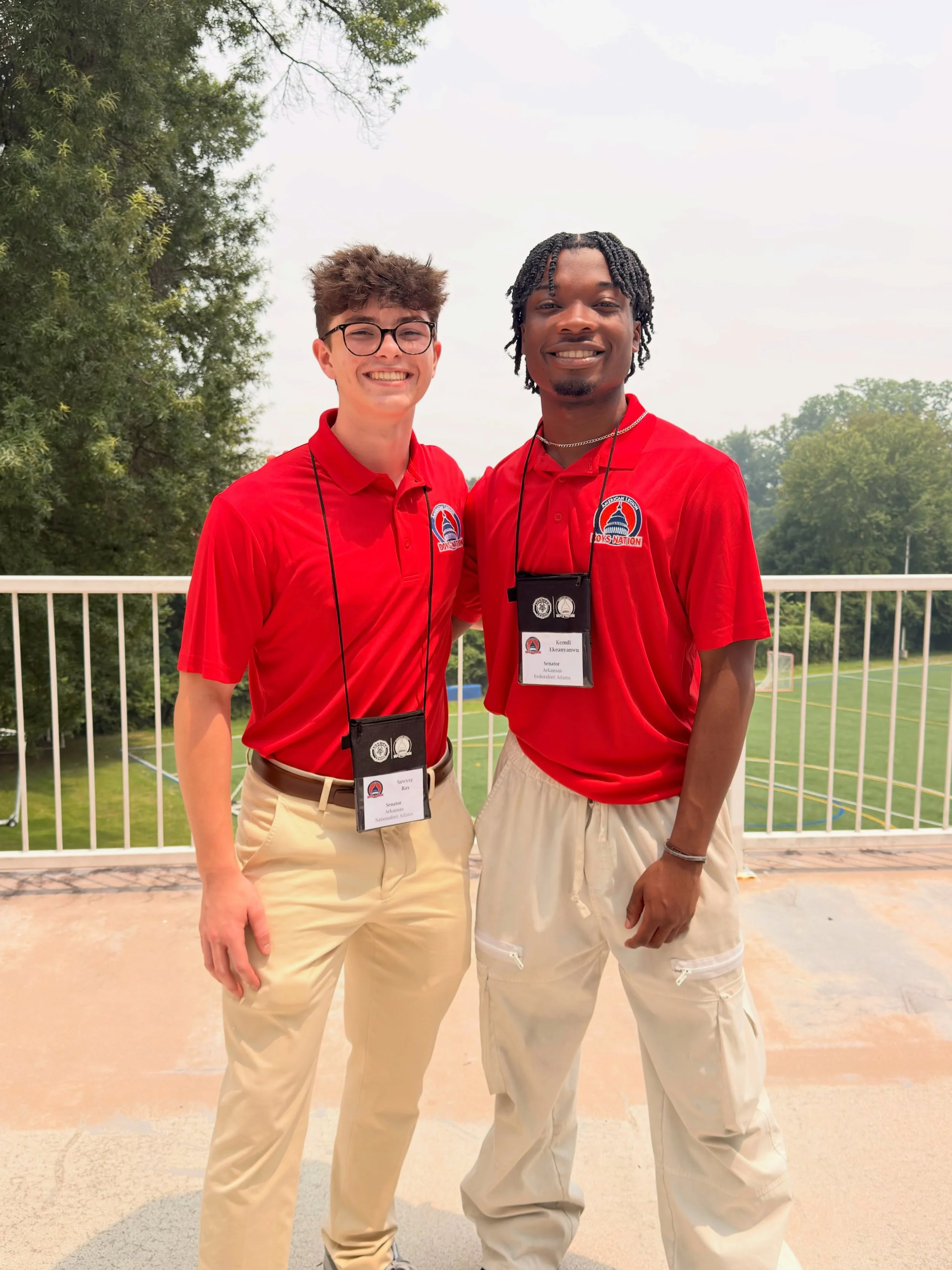 Two young men wearing red shirts with event badges around their necks, smiling and standing outdoors on a bridge, with trees and a sports field in the background.
