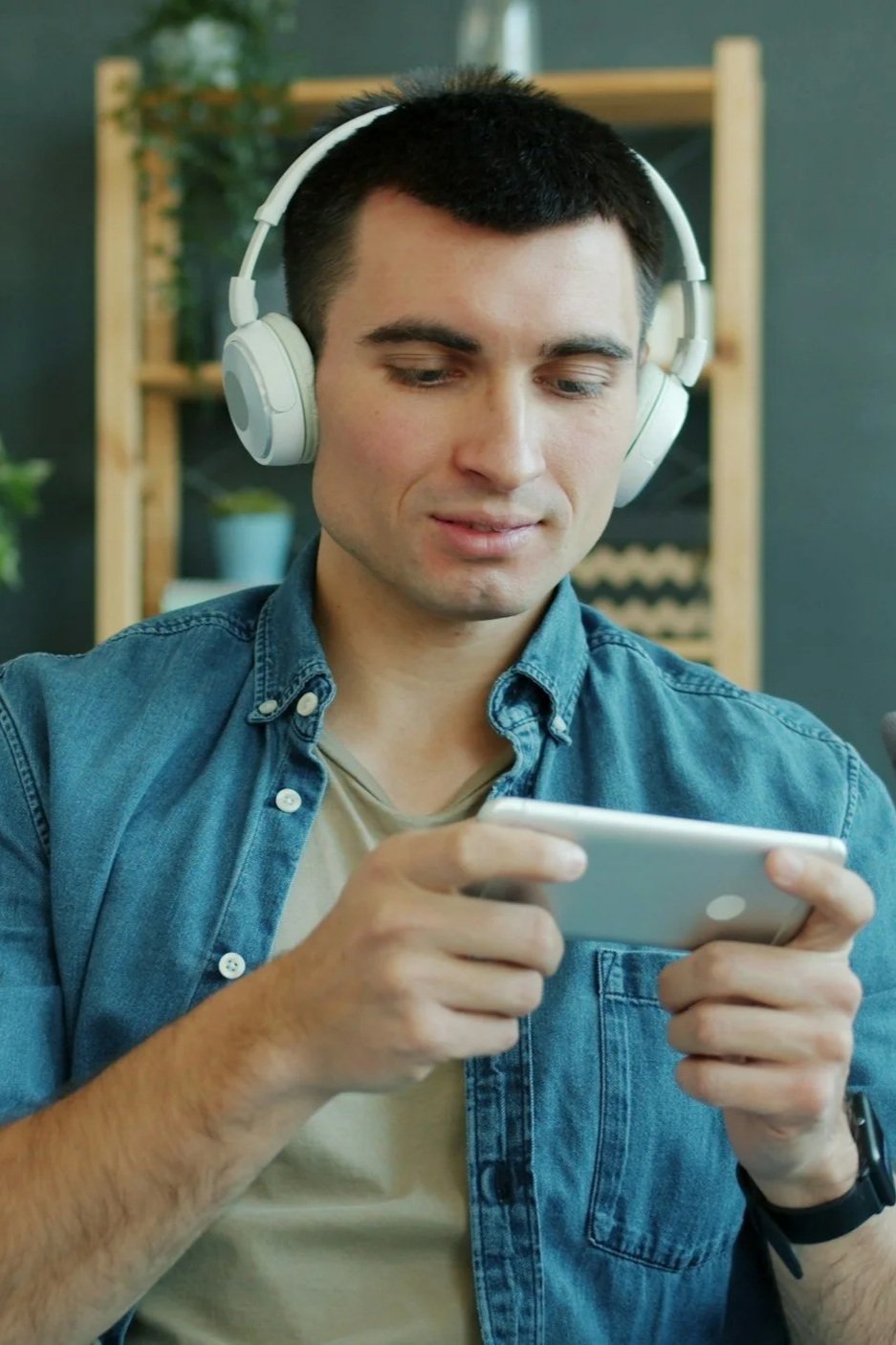 Young man wearing headphones and denim shirt, looking at a smartphone in a modern indoor setting with a plant and shelves in the background.
