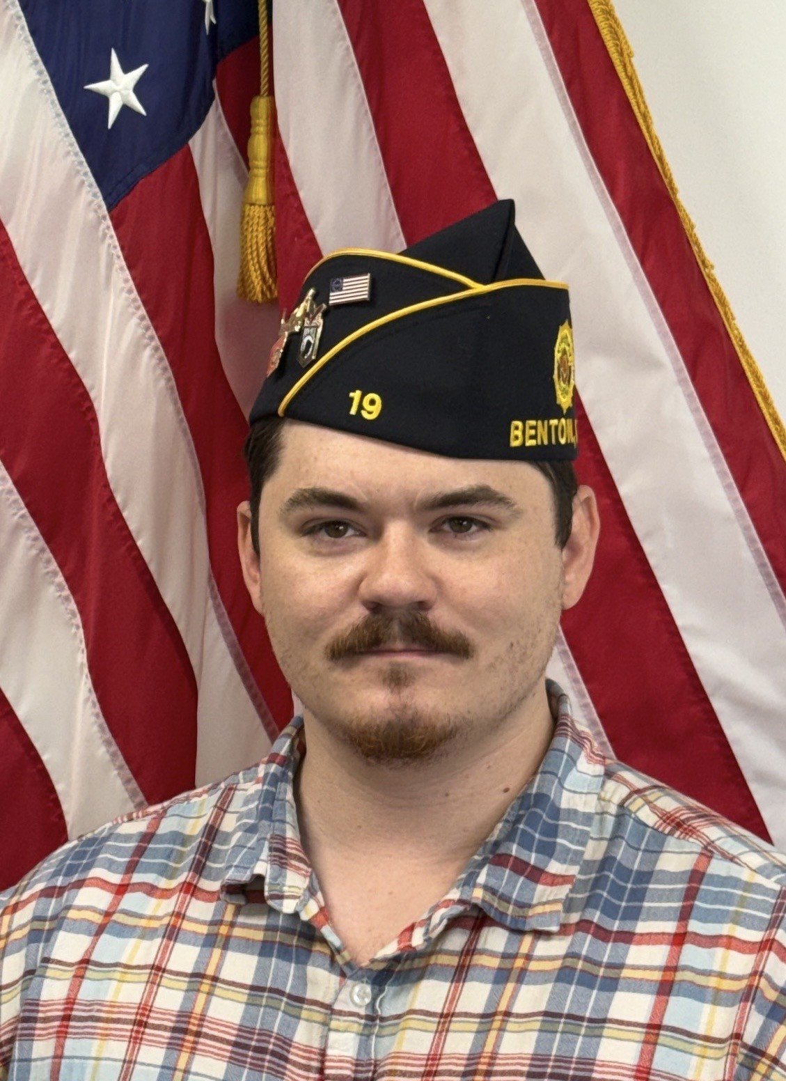 A man with a mustache and beard wearing a military-style cap with an American flag patch, standing in front of American flags.