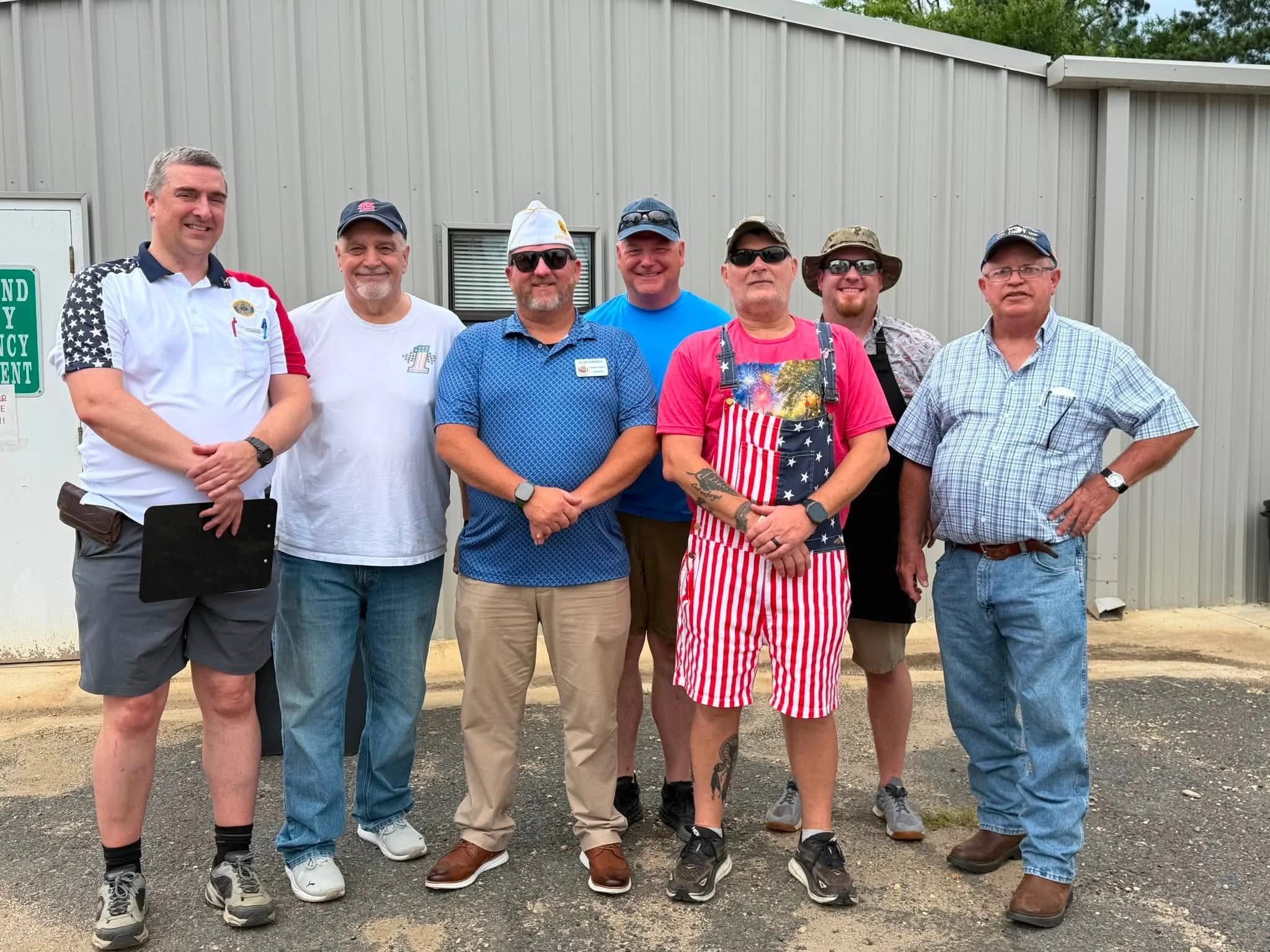 A group of eight men standing outdoors in front of a gray metal building, smiling at the camera. Some are wearing sunglasses and casual summer clothing.