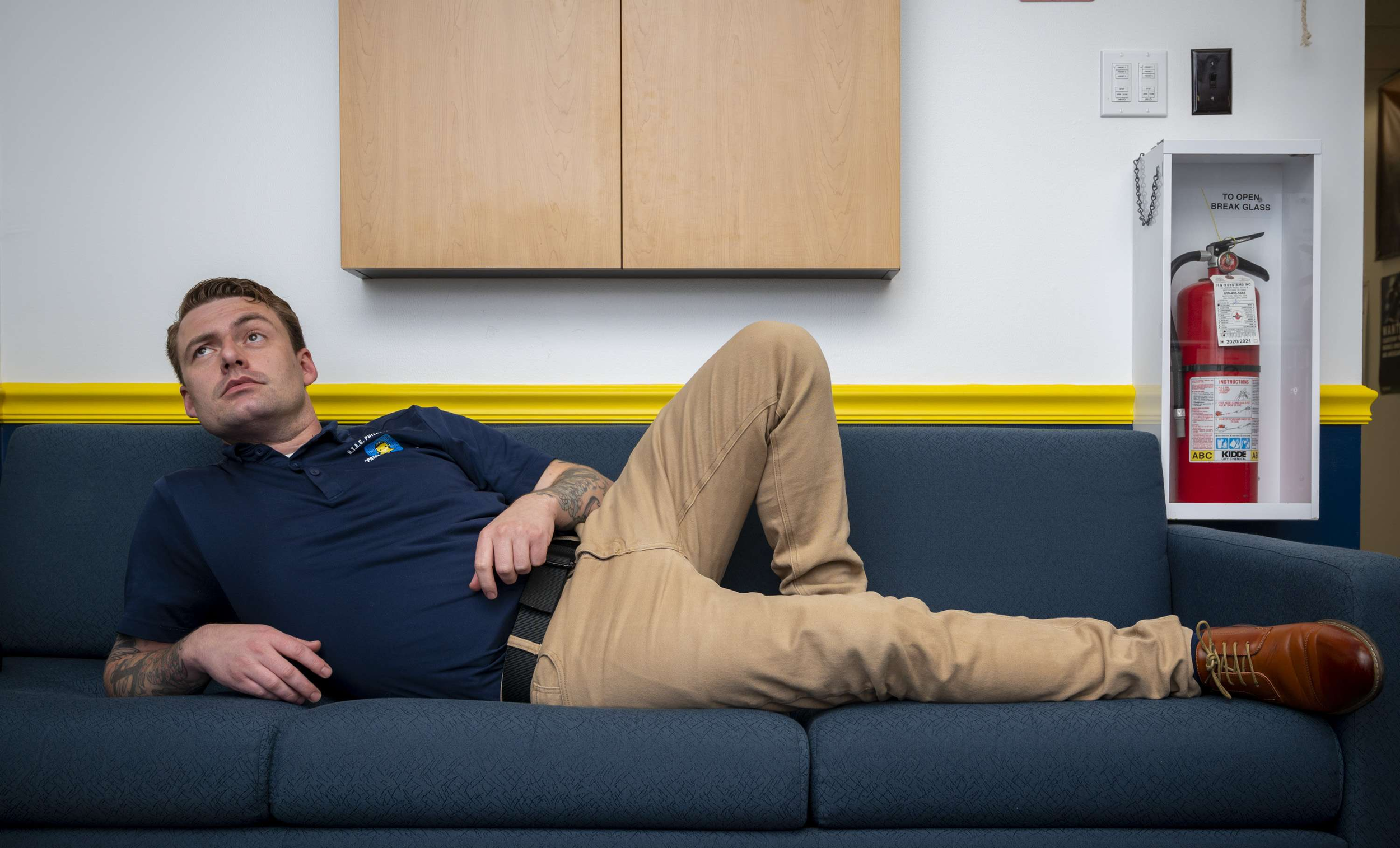 A man dressed in a navy blue polo shirt and khaki pants is reclining on a blue couch in a waiting area, looking up thoughtfully. There is a fire extinguisher in a white case mounted on the wall to his right, along with a yellow stripe running horizontally on the wall behind him.