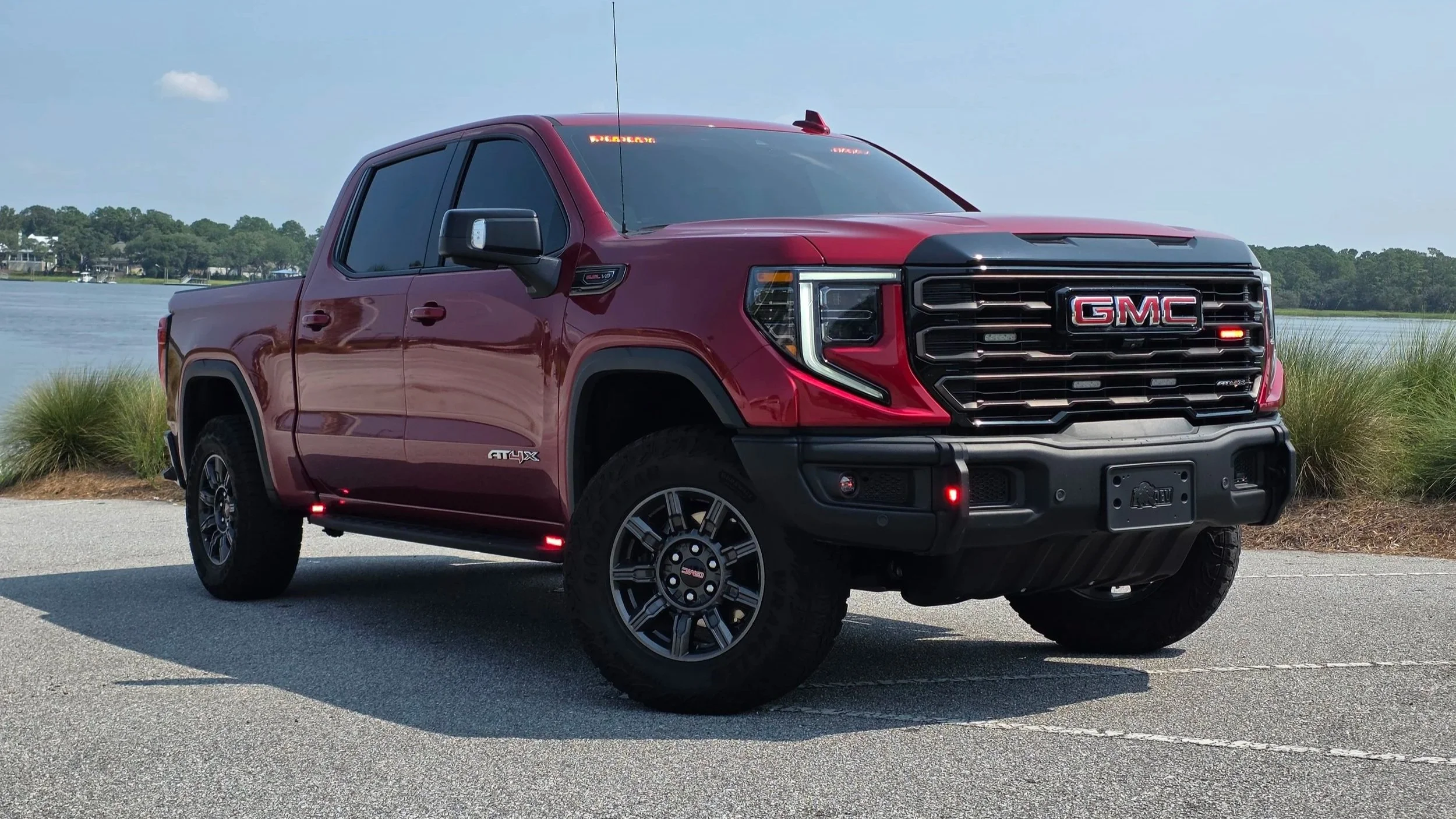 Red GMC pickup truck parked near a body of water with trees in the background under a partly cloudy sky.