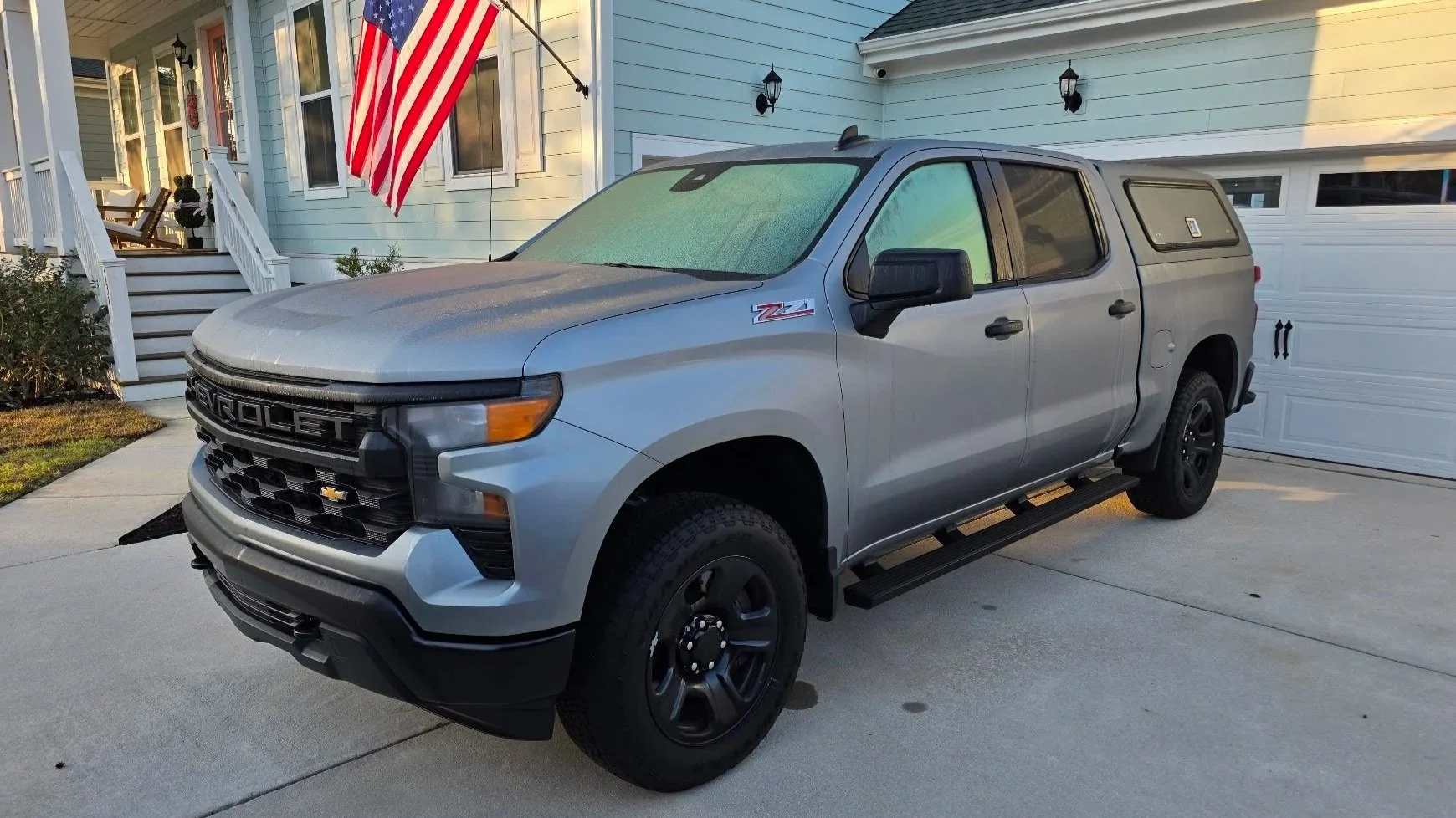 Silver Chevrolet Silverado pickup truck parked in driveway in front of a house with light blue siding, white porch, American flag, and white garage door.