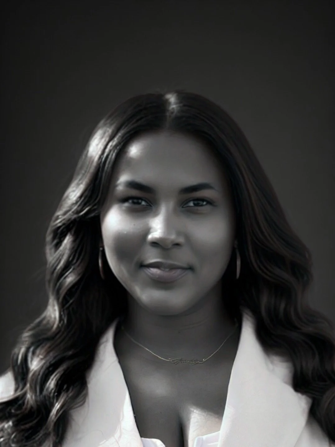 Black and white portrait of a woman with long wavy hair, wearing earrings, a necklace, and a light-colored top, with a dark background.