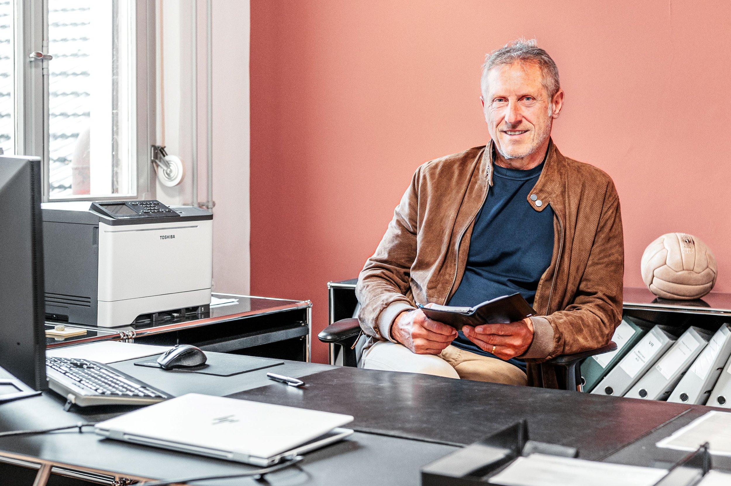 Ein älterer Mann sitzt an einem Büro schreibtisch, hält ein Notizbuch in den Händen und lächelt. Im Hintergrund ist eine rote Wand, und auf einem Regal steht ein Volleyball. Das Büro ist mit einem Computer, einem Drucker und anderen Büromaterialien ausgestattet.