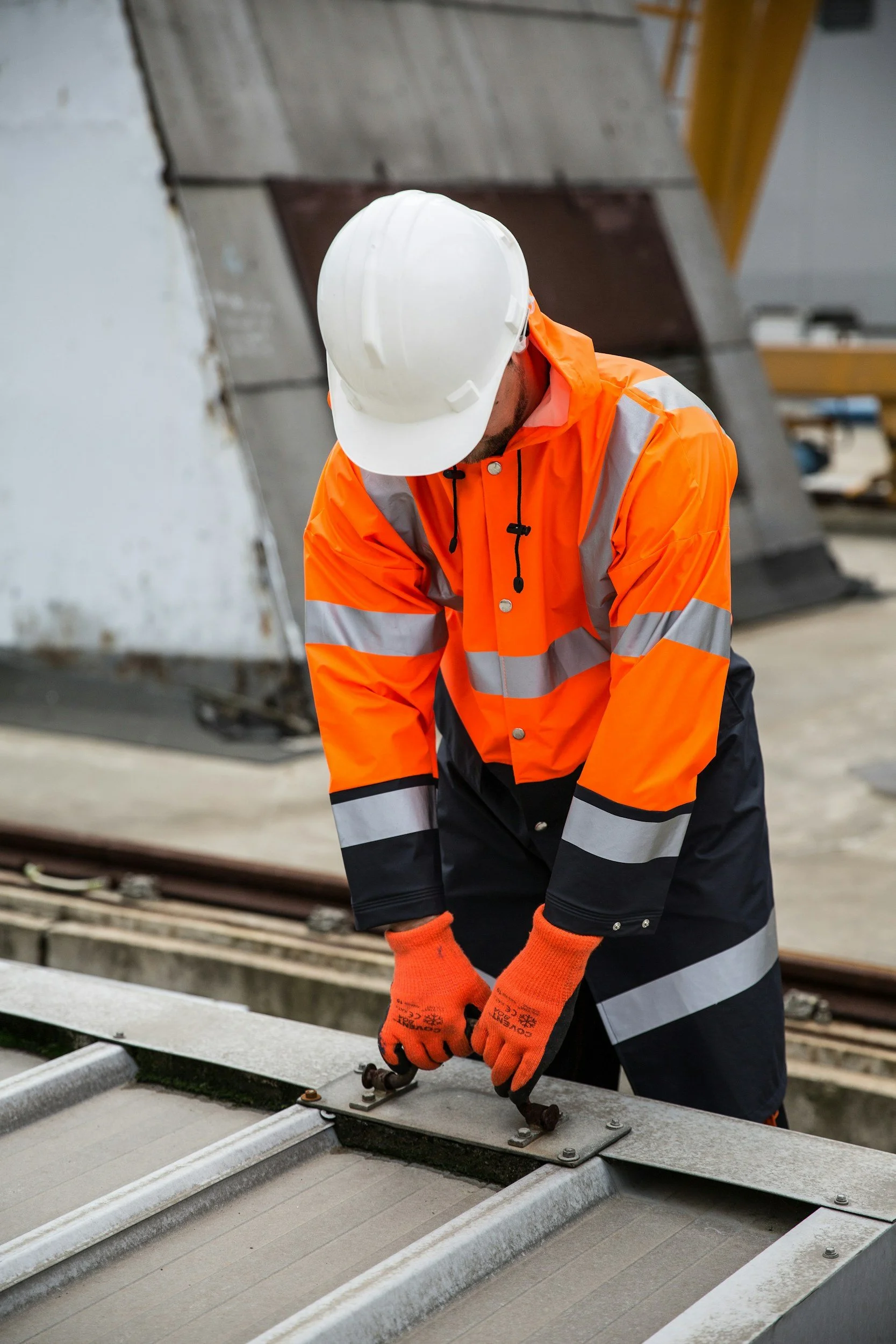 Ein Bauarbeiter trägt eine weiße Schutzmütze, eine orangeWarnweste mit reflektierenden Streifen, orange Handschuhe und arbeitet an einer Baustelle mit Metall- und Holzstrukturen.