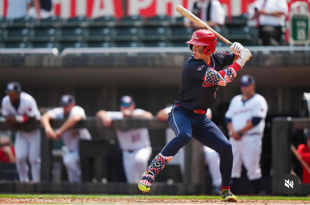 A baseball player in a patriotic uniform at bat during a game, with teammates and coaches in the background.