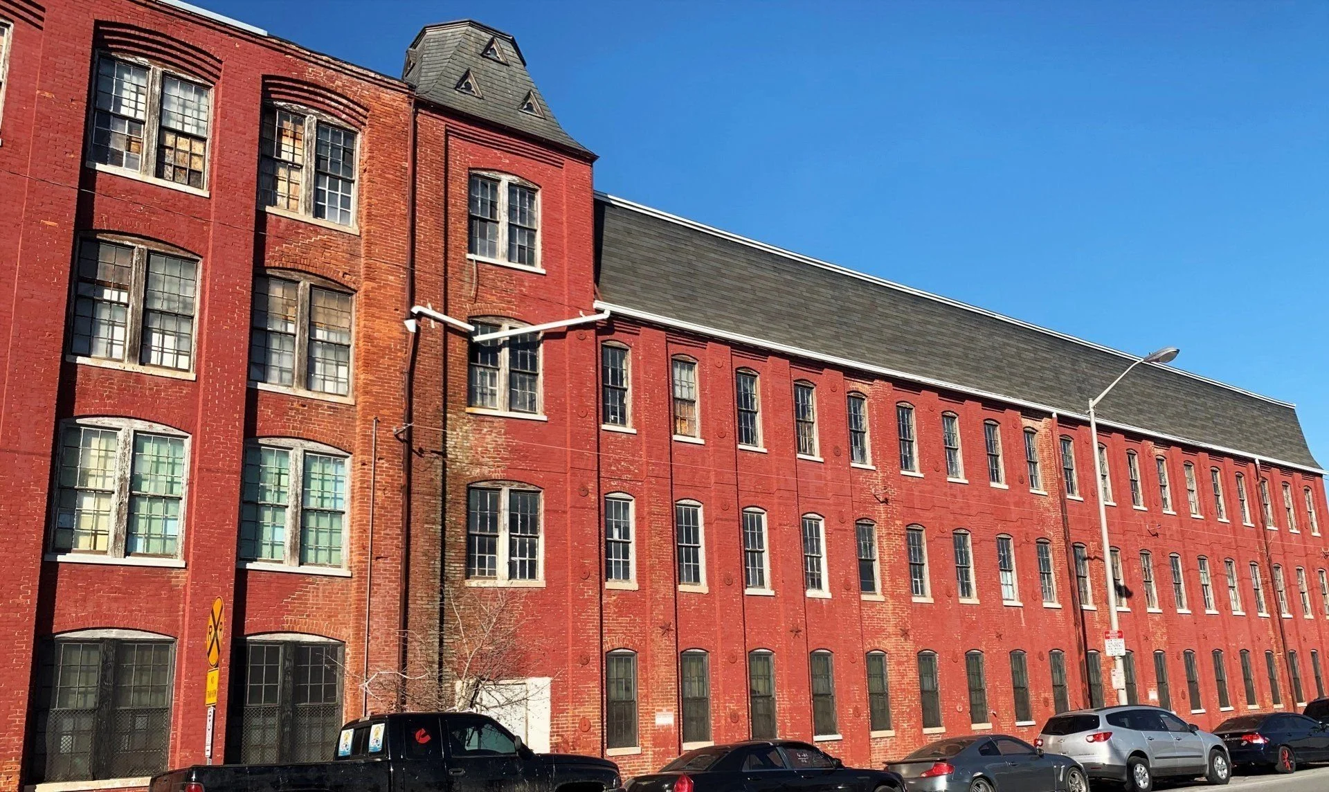 A tall red brick building with multiple windows and a dark gray sloped roof, set against a clear blue sky. There are cars parked along the street in front of the building, and a streetlamp is visible.