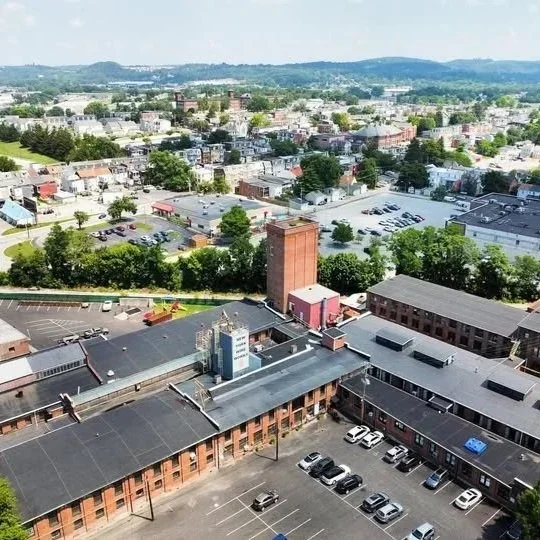 Aerial view of a city with buildings, parking lots, and streets, surrounded by trees and hills in the background.