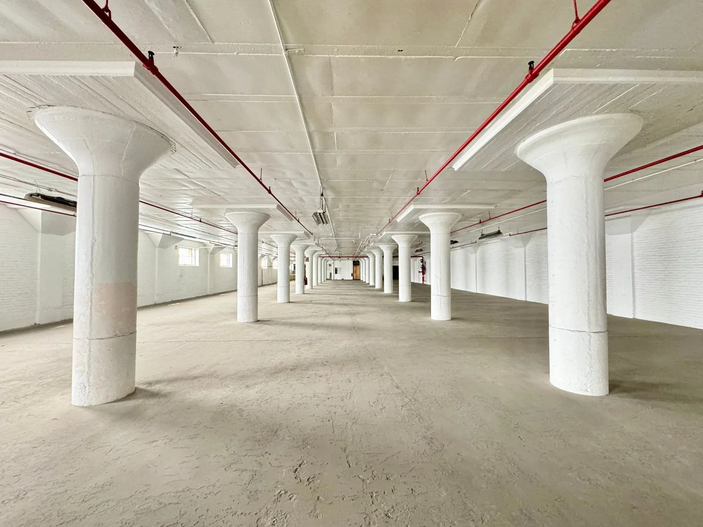 Empty white-walled parking garage with columns, concrete floor, and exposed ceiling with red pipes.