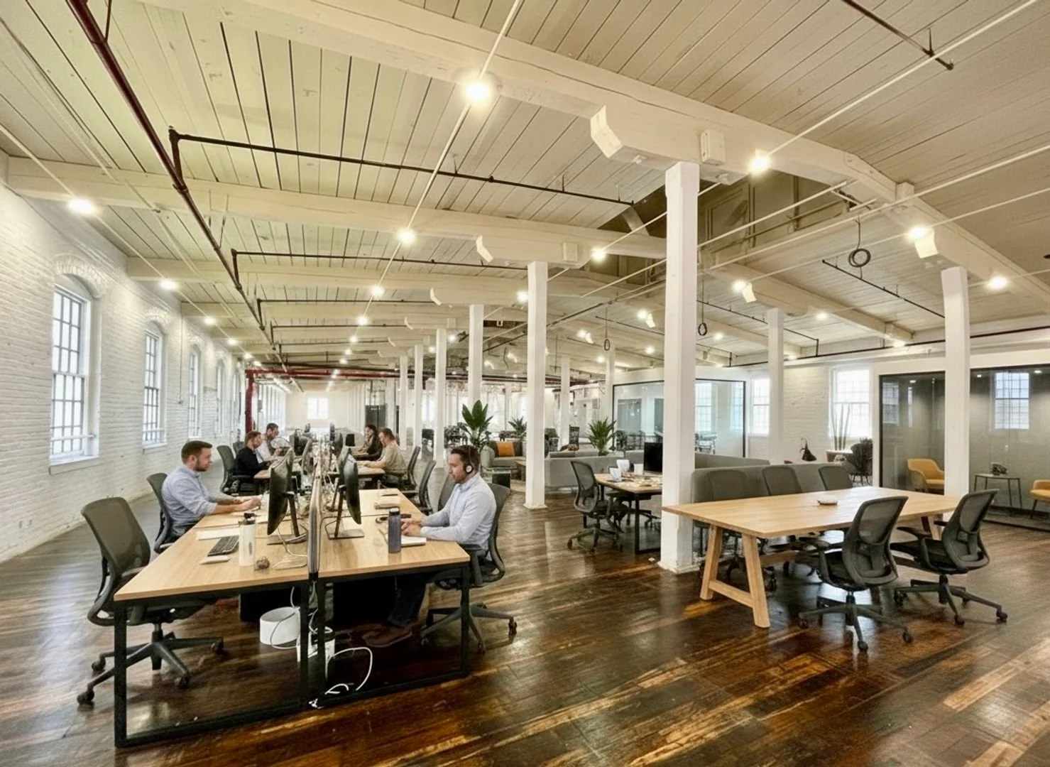 Open-plan office with employees working at desks with computers, large windows, exposed ceilings, and a polished wooden floor.