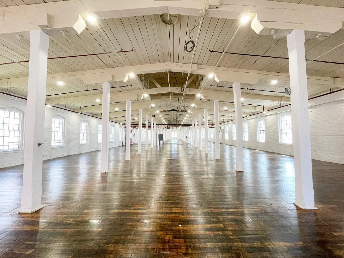 Spacious, empty industrial-style room with white brick walls, large windows, wooden flooring, white support columns, and a high ceiling with exposed lighting fixtures.