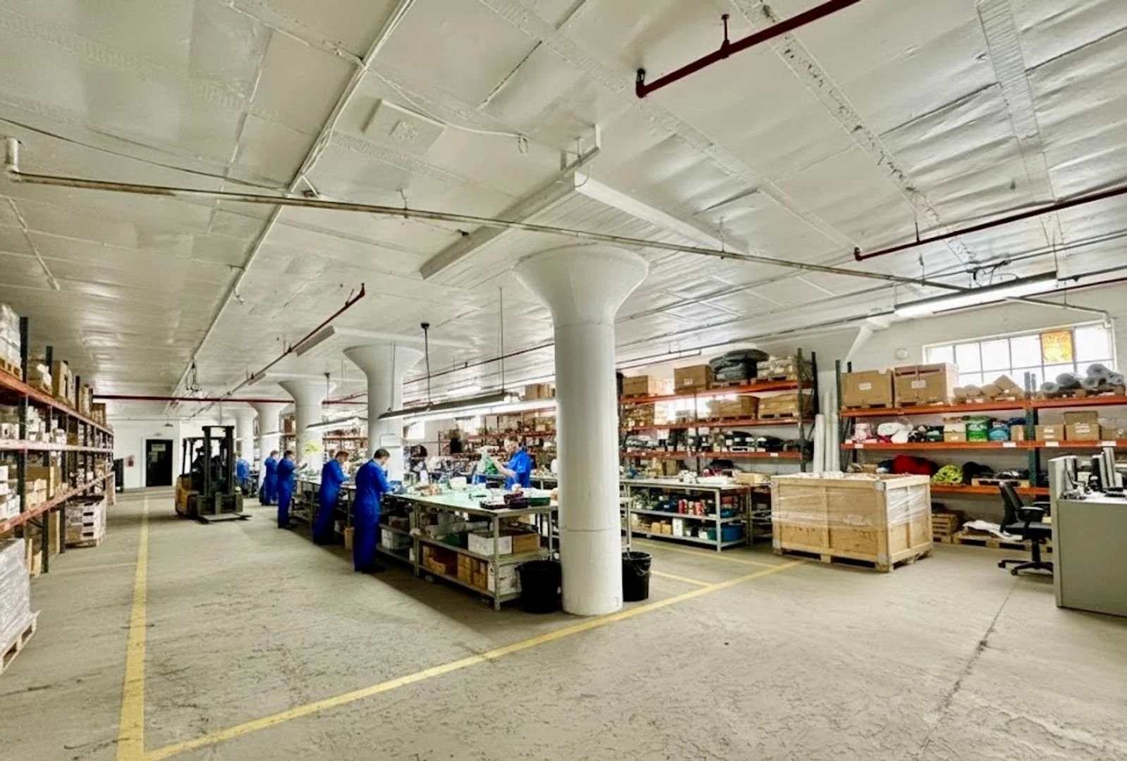 Warehouse with workers in blue uniforms working at workbenches amidst shelves filled with boxes and supplies, concrete floor, large white columns, high ceiling with pipes and lighting.