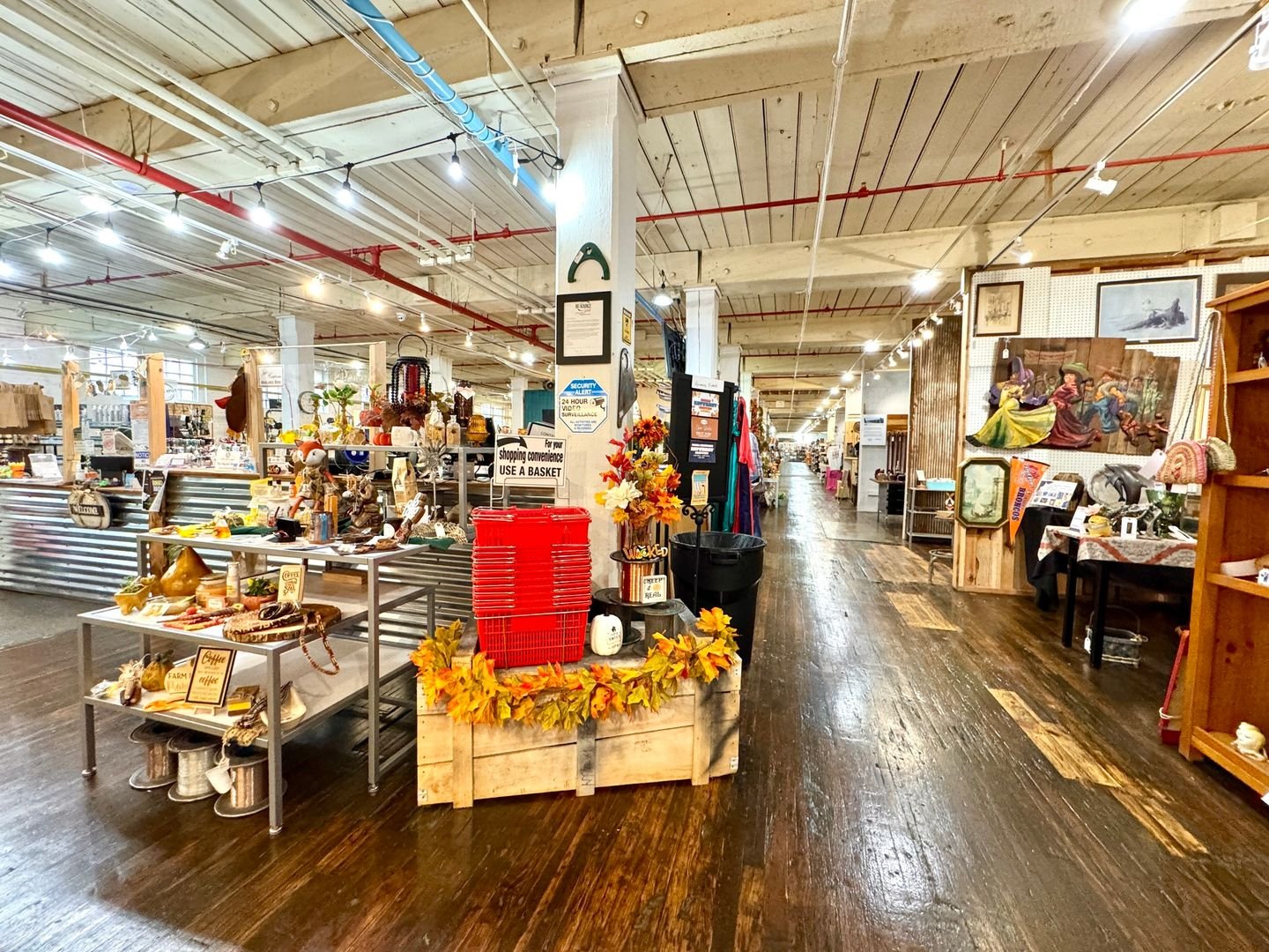 Interior of a thrift or antique store with various vintage items, decorations, and furniture displayed on tables and shelves, with a wooden floor, a high ceiling, and warm lighting.