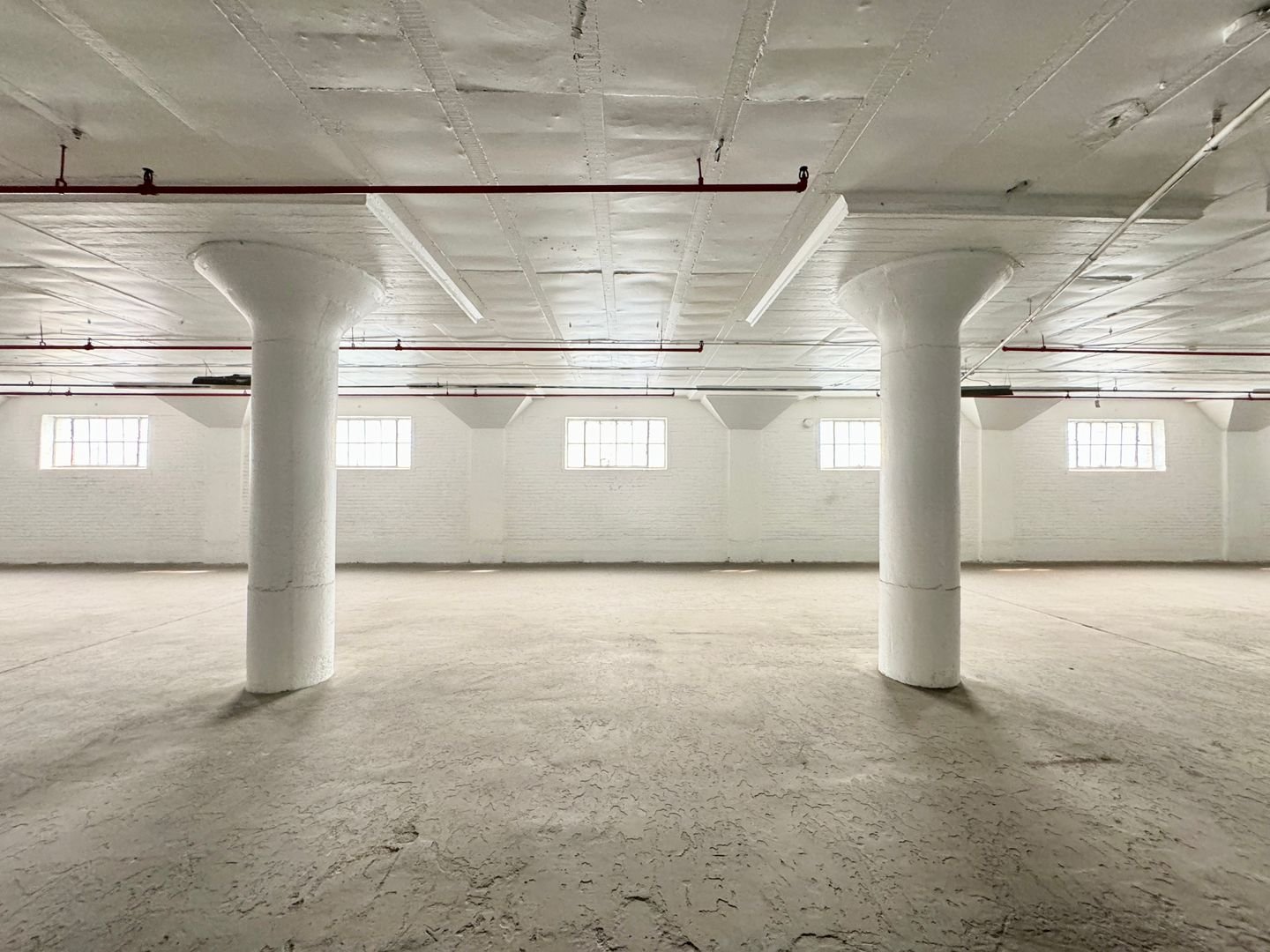 Empty industrial space with white painted brick walls, concrete floor, and large windows, supported by several white columns.