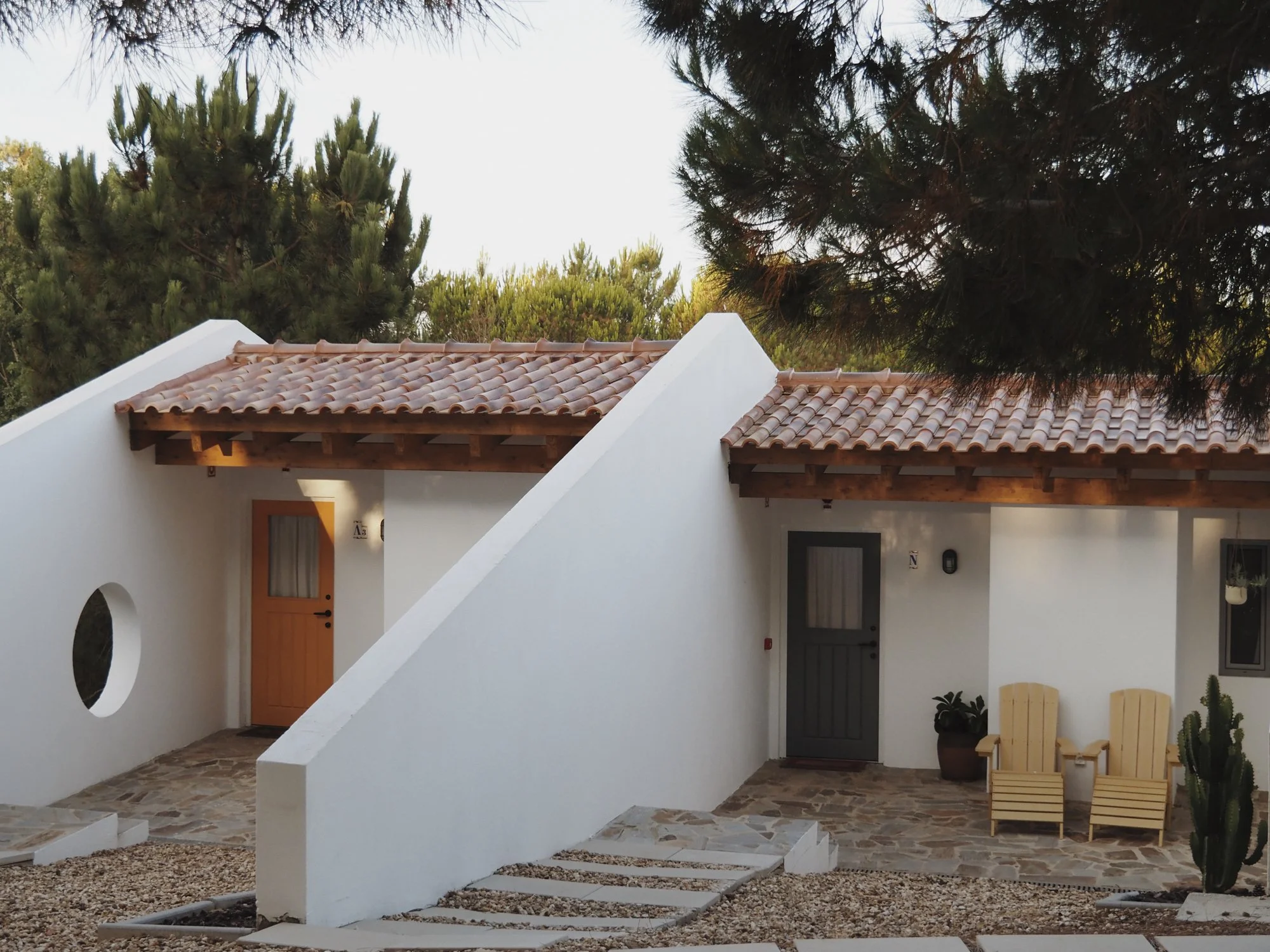 Hillside Quarto, modern house with terracotta tiled roof, surrounded by greenery and outdoor seating including wooden chairs and potted cactus plants.