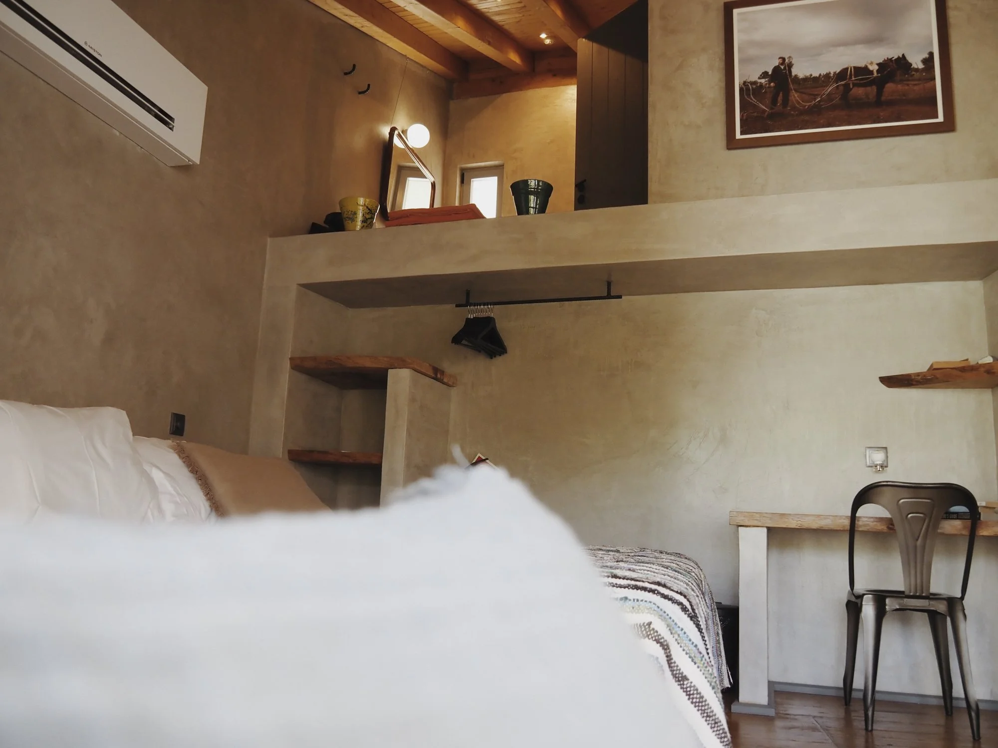 A cozy, warmly lit room with beige walls and wooden accents, featuring a bed with a white and brown striped blanket, a wooden chair, a framed horse photograph, small windows, and a decorative mirror.
