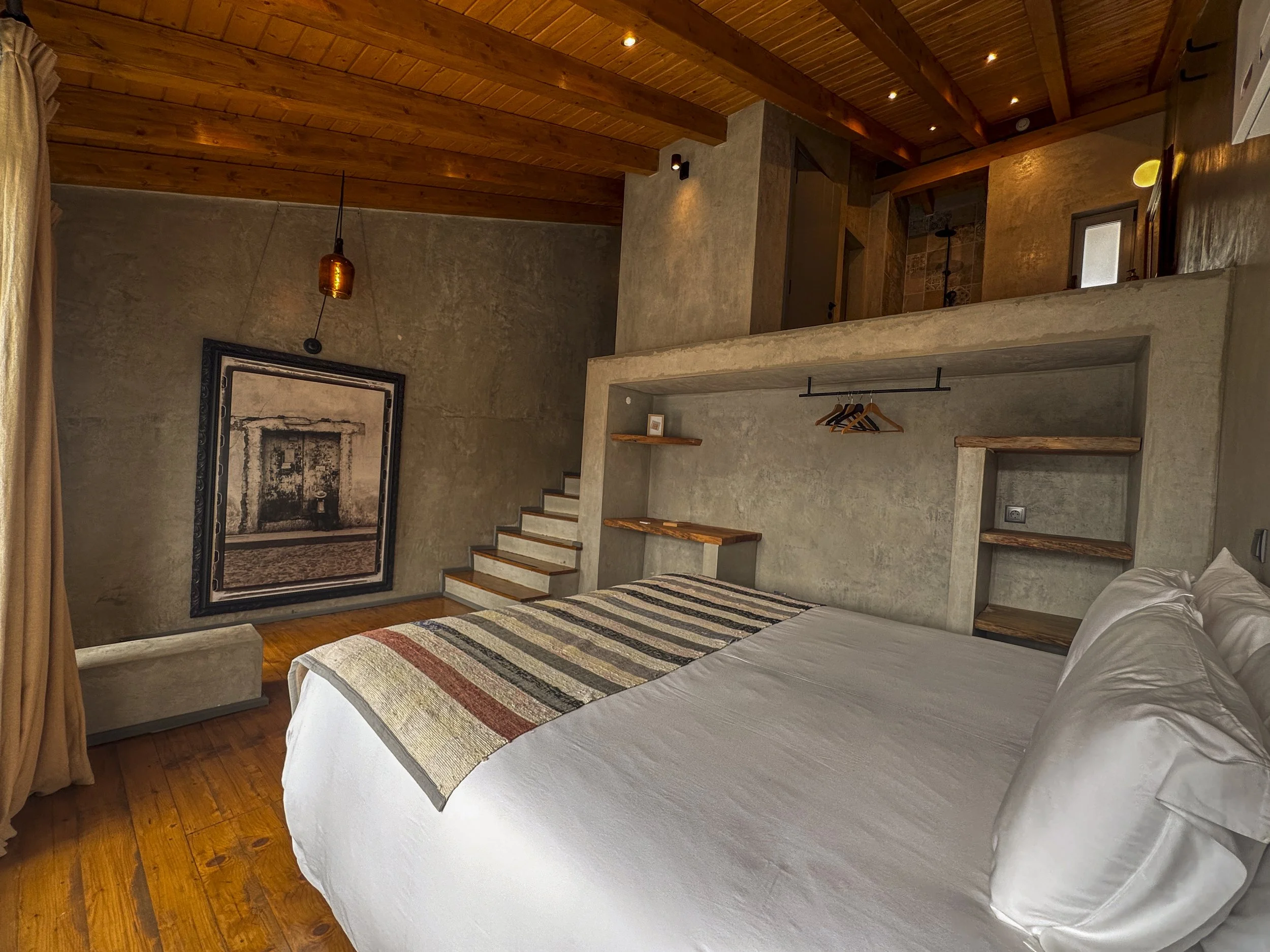 A bedroom with a bed covered with a striped quilt, a painting on the wall, a hanging light, and wooden beams on the ceiling.