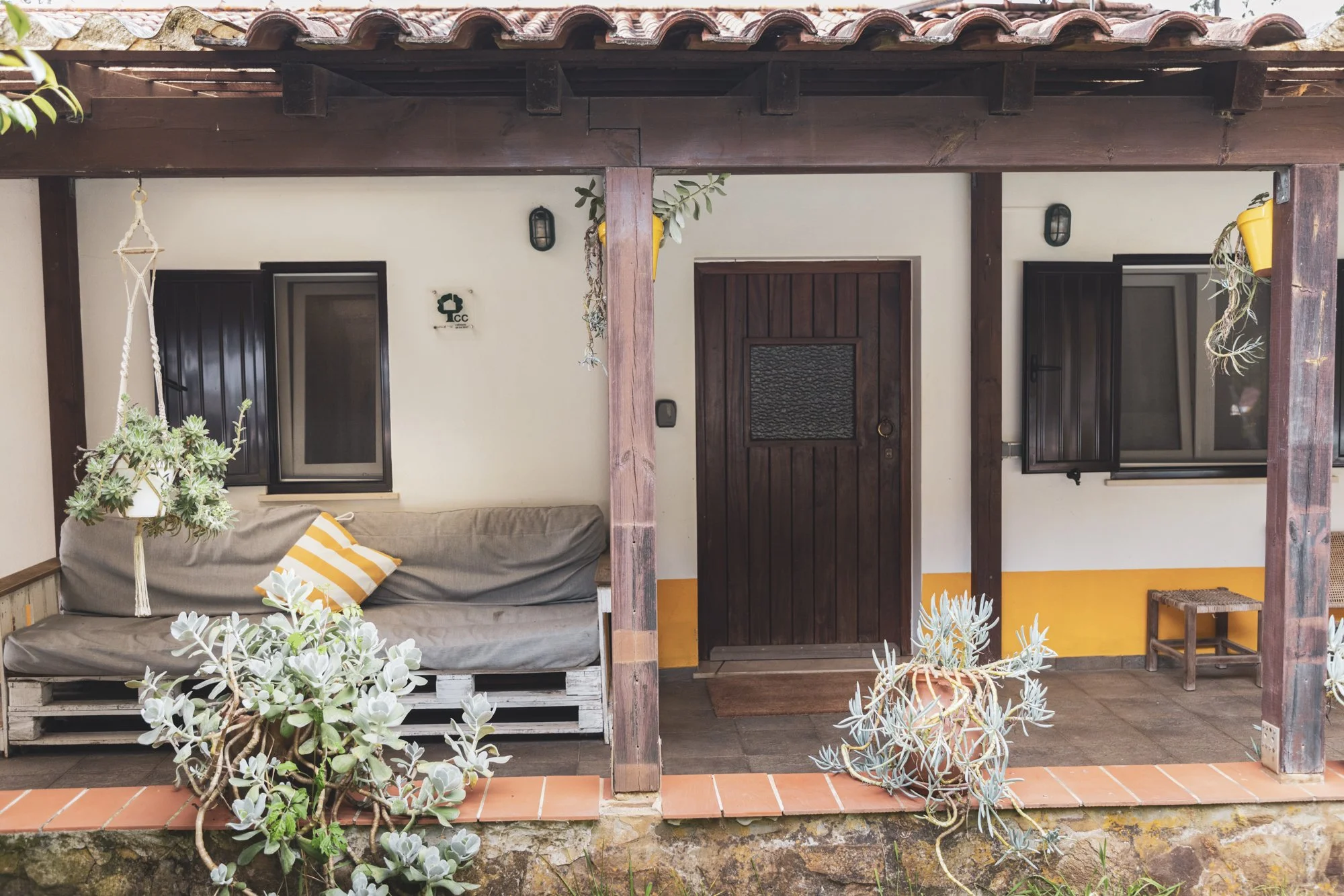 A cozy outdoor patio area with a wooden pergola, a dark wood door, two black window frames, and potted succulents. The space includes a beige sofa with a striped pillow, hanging plants, and string lights.