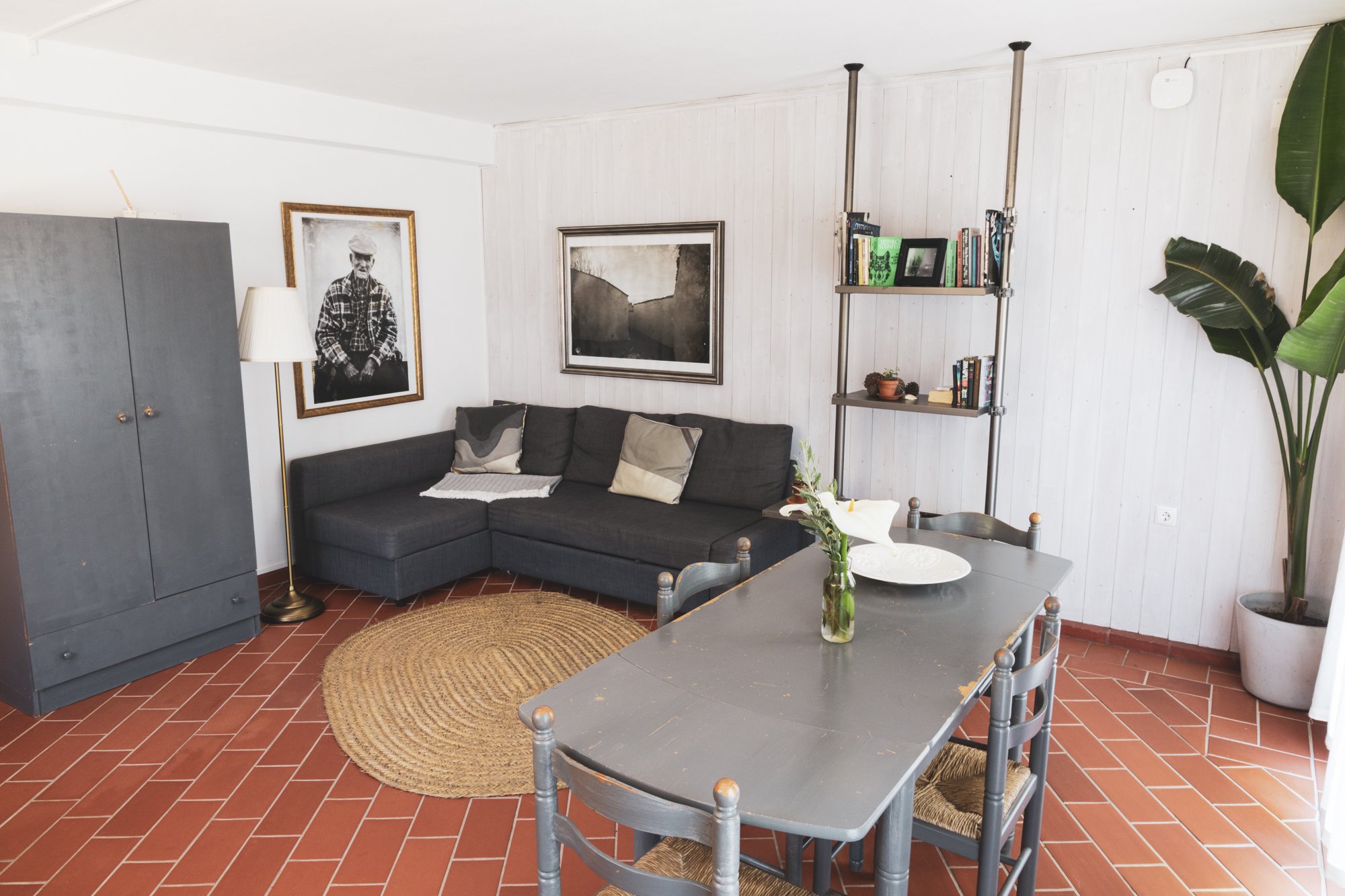 A cozy living room with a dark gray sectional sofa holding beige and patterned pillows, a round woven rug on terracotta tiled flooring, a gray wardrobe, framed black and white photographs on white paneled walls, a metal bookshelf with books and decorative items, a tall potted plant, and a dining table with a vase of white flowers.