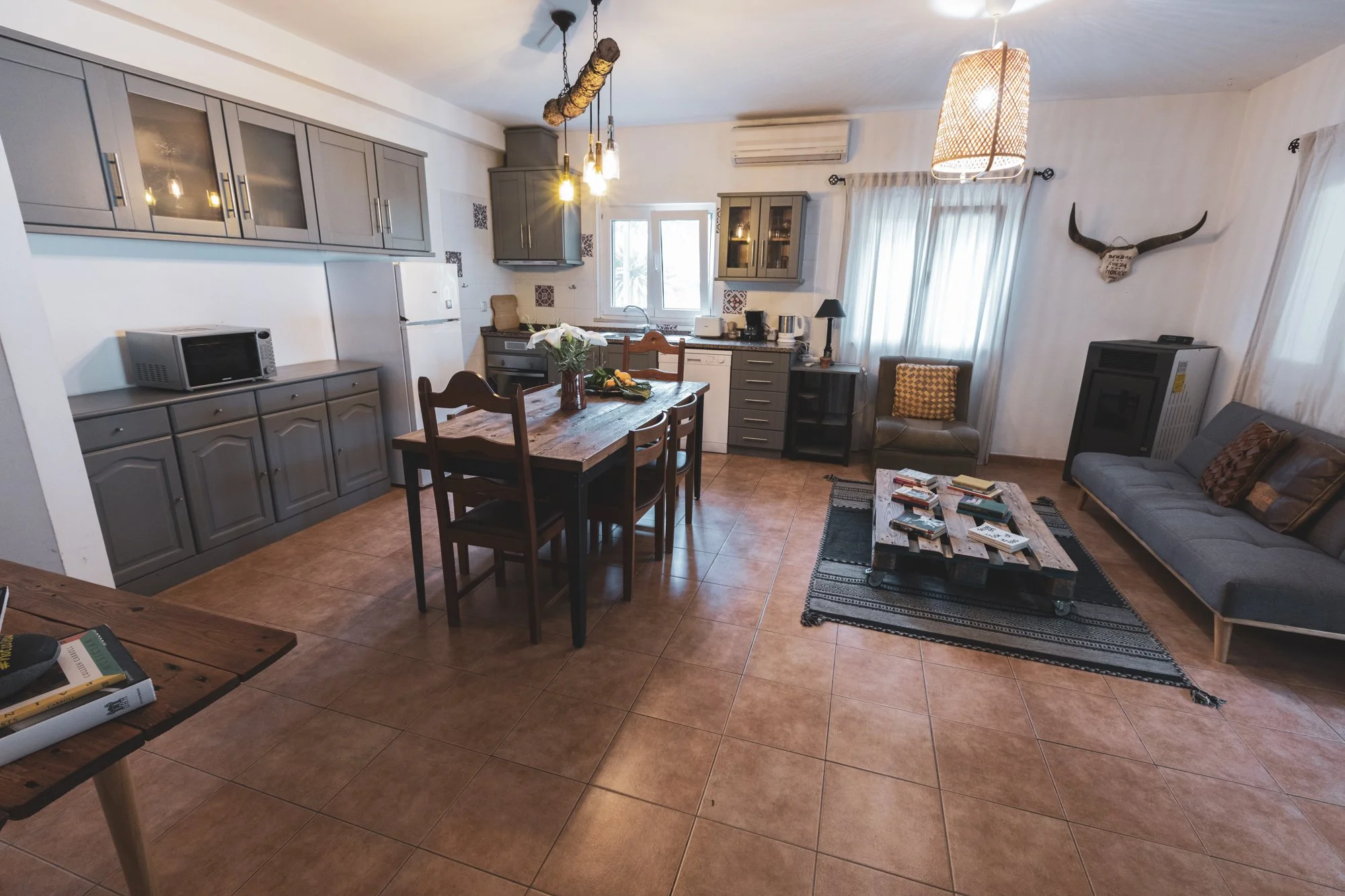 A cozy living room and kitchen area in Casa Mae, with tile flooring, a wooden dining table with chairs, a gray sofa, a coffee table with books, and various decorative items.