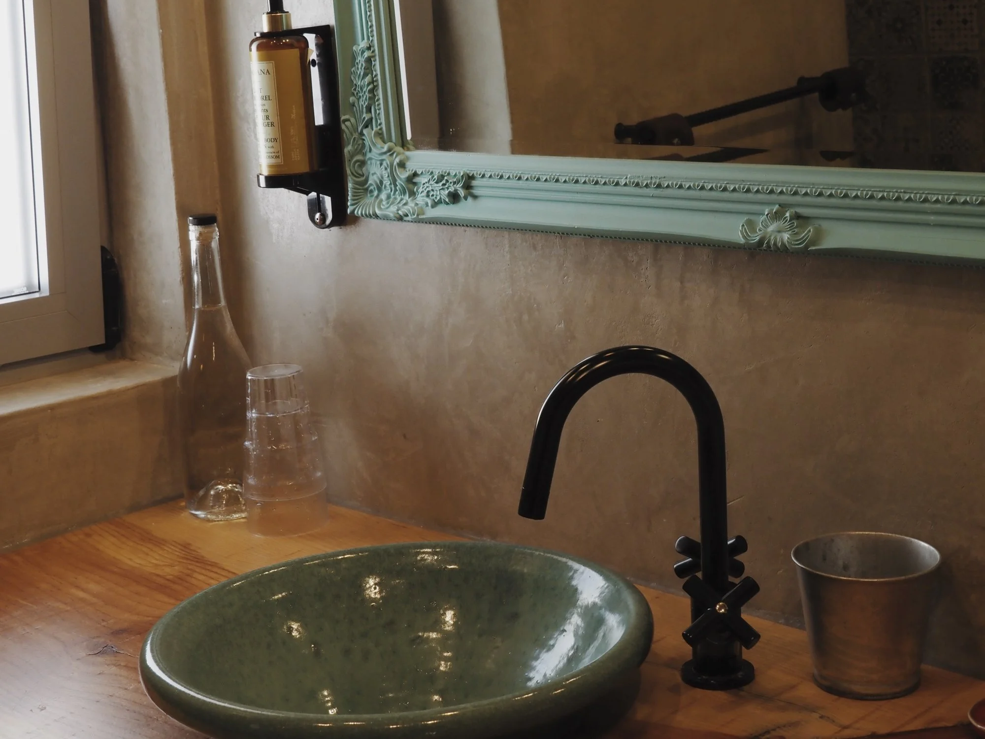 Green ceramic sink, black faucet, clear glass cup, soap dispenser, mirror with ornate turquoise frame, window ledge with a bottle and glass.