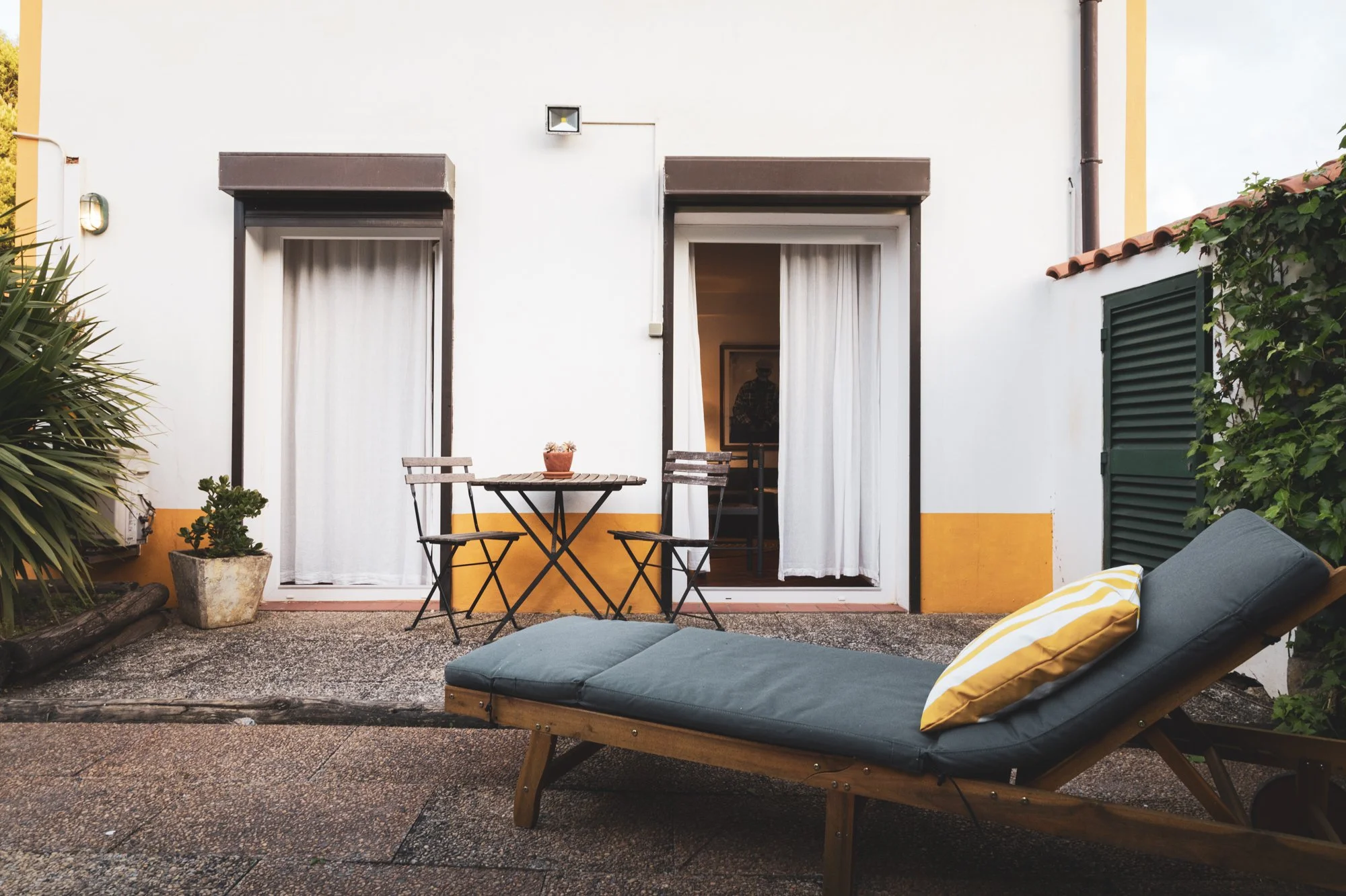 A cozy outdoor patio with a small round metal table and two chairs, decorated with a potted plant, and a large cushioned lounge chair with yellow-striped pillows, located in front of white house walls with brown trim and curtains, and surrounded by greenery.
