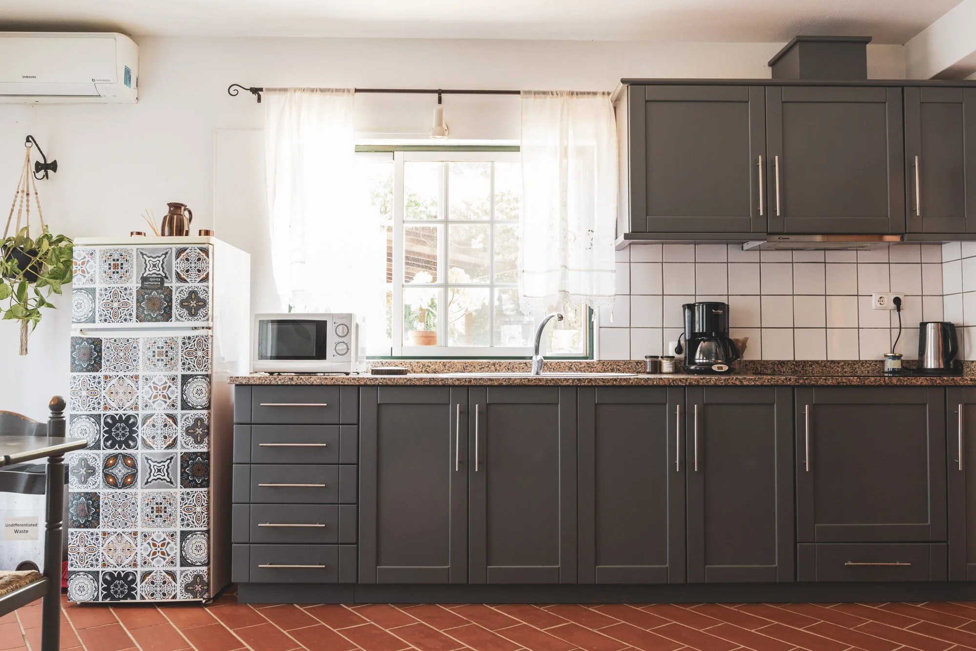 A kitchen with gray cabinets, a window with sheer curtains, a microwave, a coffee maker, and a refrigerator with decorative tiles on its side.
