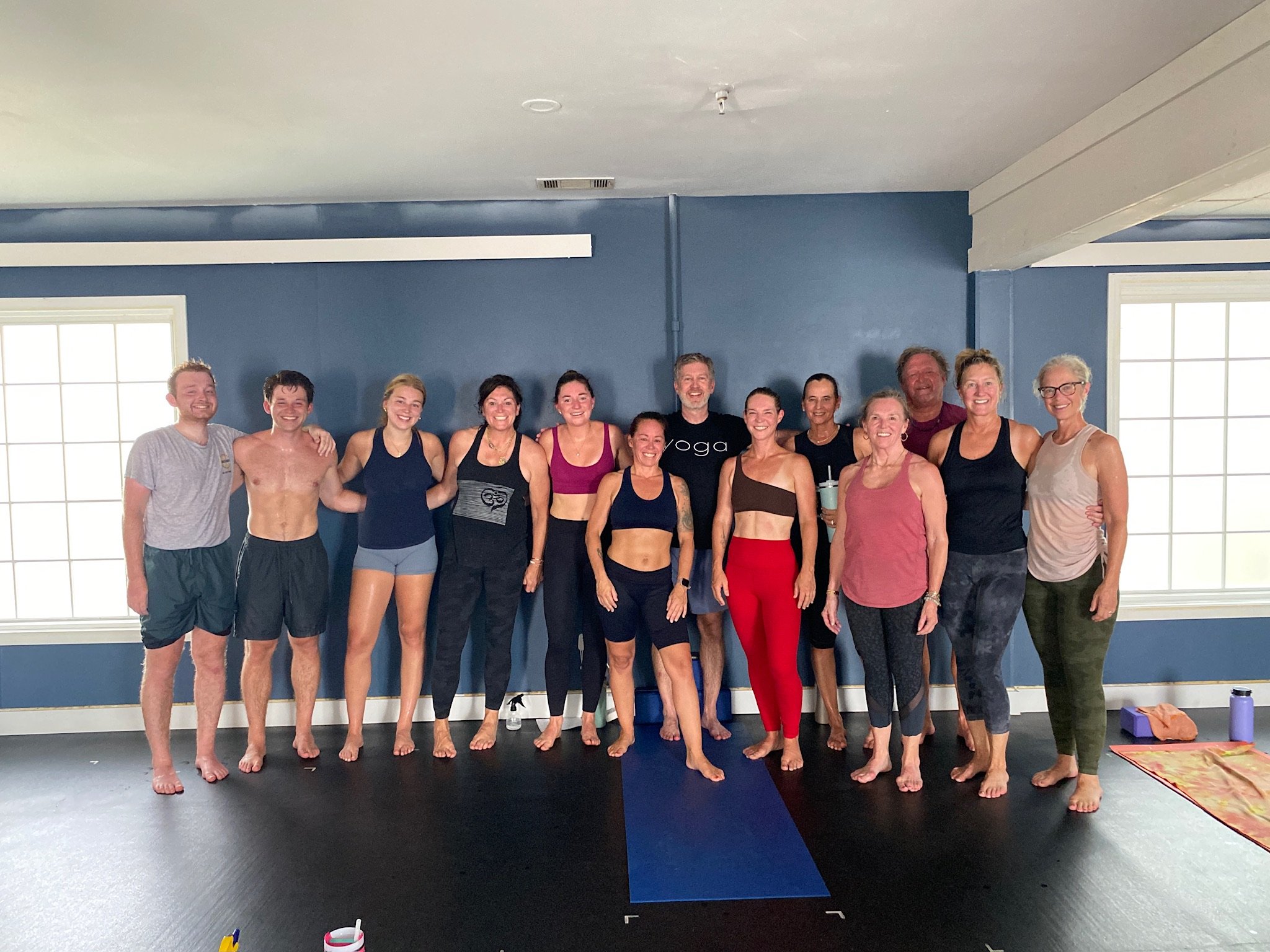 Group of people standing together in a yoga studio, smiling at the camera, with some wearing athletic clothing and others in casual attire.