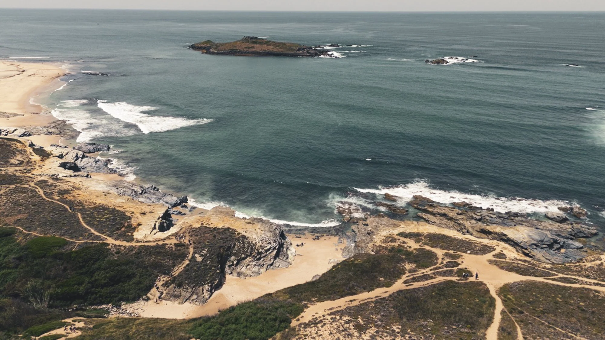 Aerial view of rocky Portugal coastline with sandy beach, ocean waves, and small island in the distance.