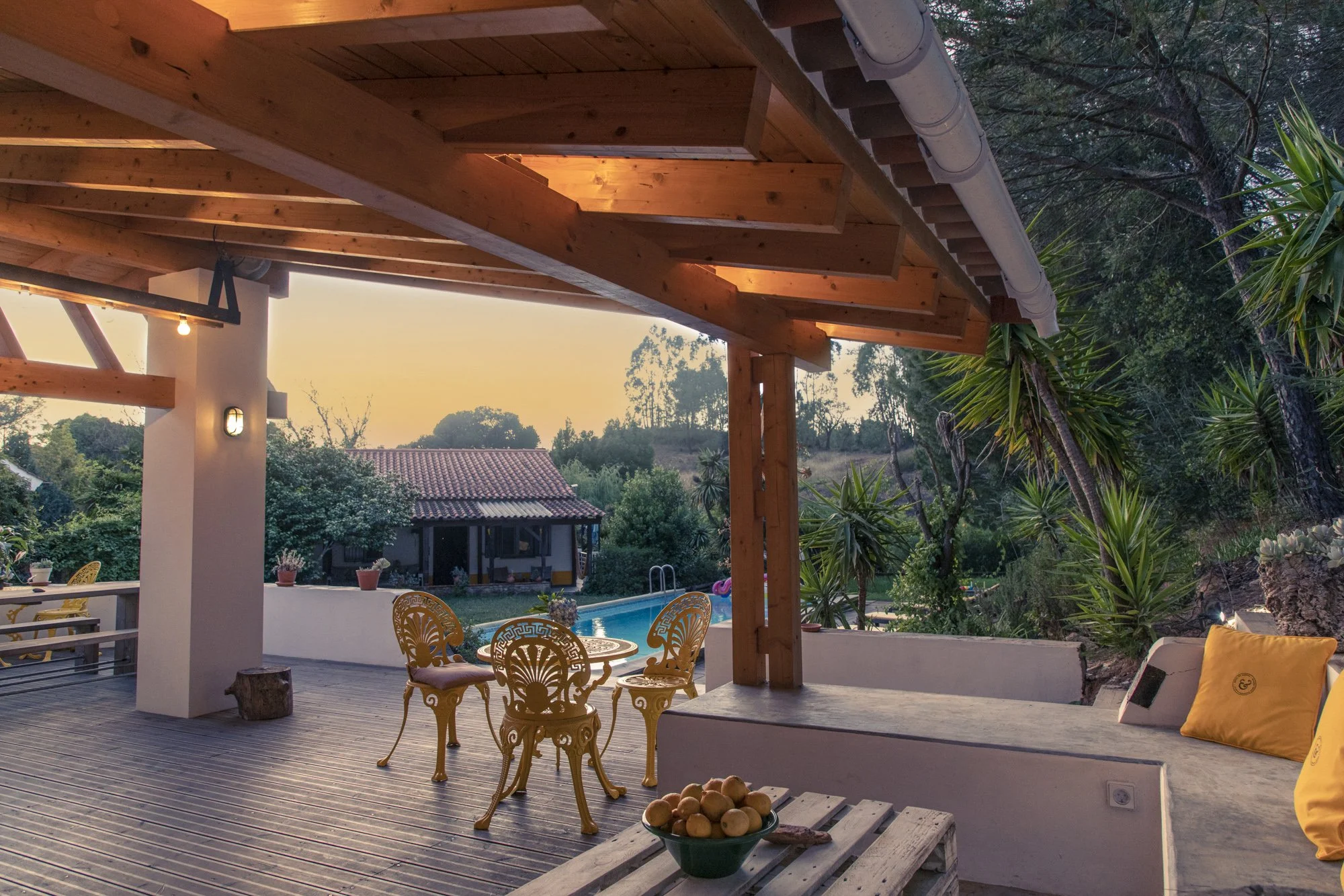 Outdoor patio area with wooden deck, yellow ornate metal chairs around a matching table, yellow cushions, a bowl of apples, and string lights above. In the background, a swimming pool and lush greenery with trees and a small house with a tiled roof are visible at sunset.