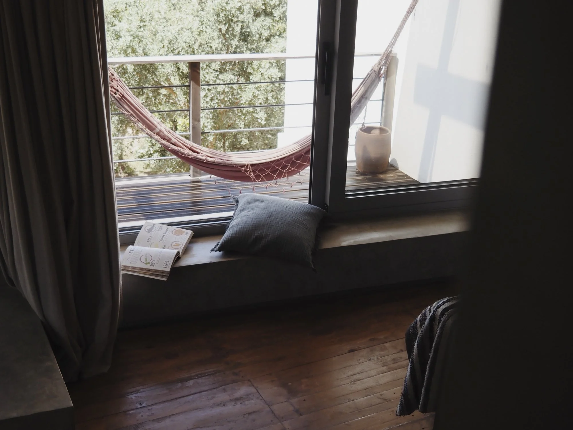 View of a balcony seen through an open sliding glass door, with a pink hammock, a potted plant, a pillow, and a newspaper on the window sill, overlooking green trees outside.