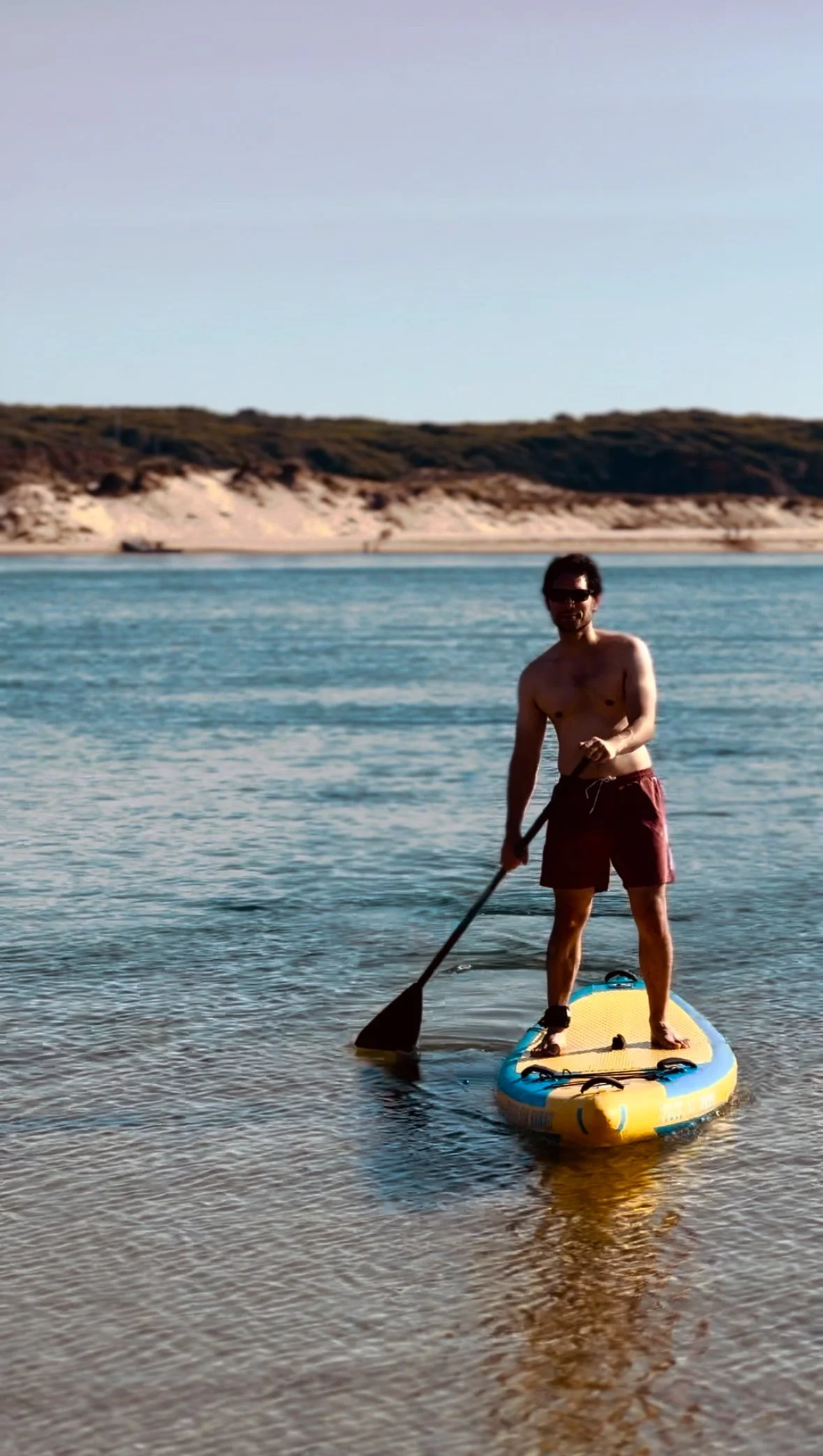 A person standing on a paddleboard in Portugal yoga retreat shallow water, wearing sunglasses and red shorts, holding a paddle, with a background of sandy cliffs and a clear sky.