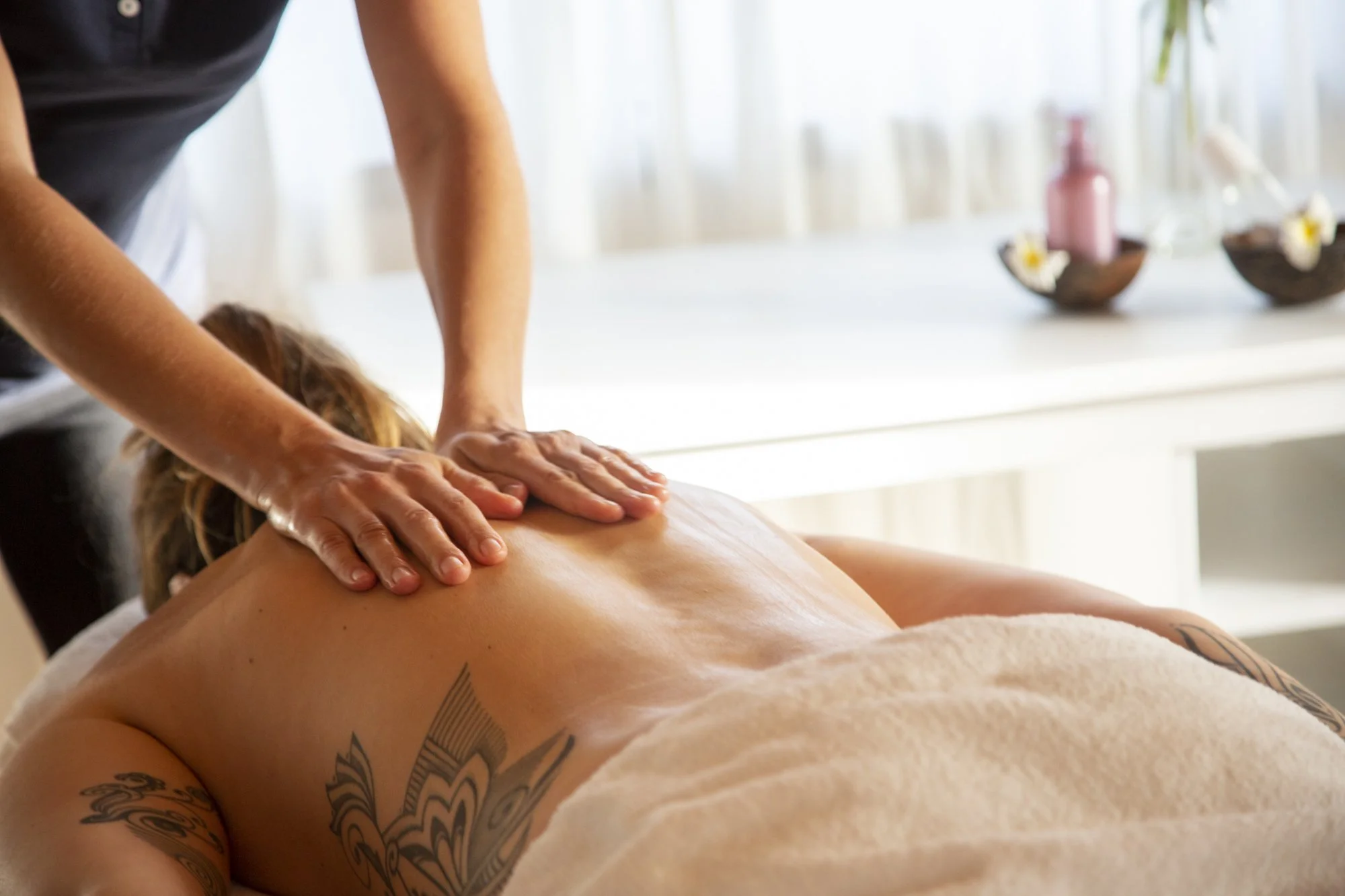 A person receiving a massage on their back in a bright, relaxing spa setting with decorative bowls and a pink bottle in the background.