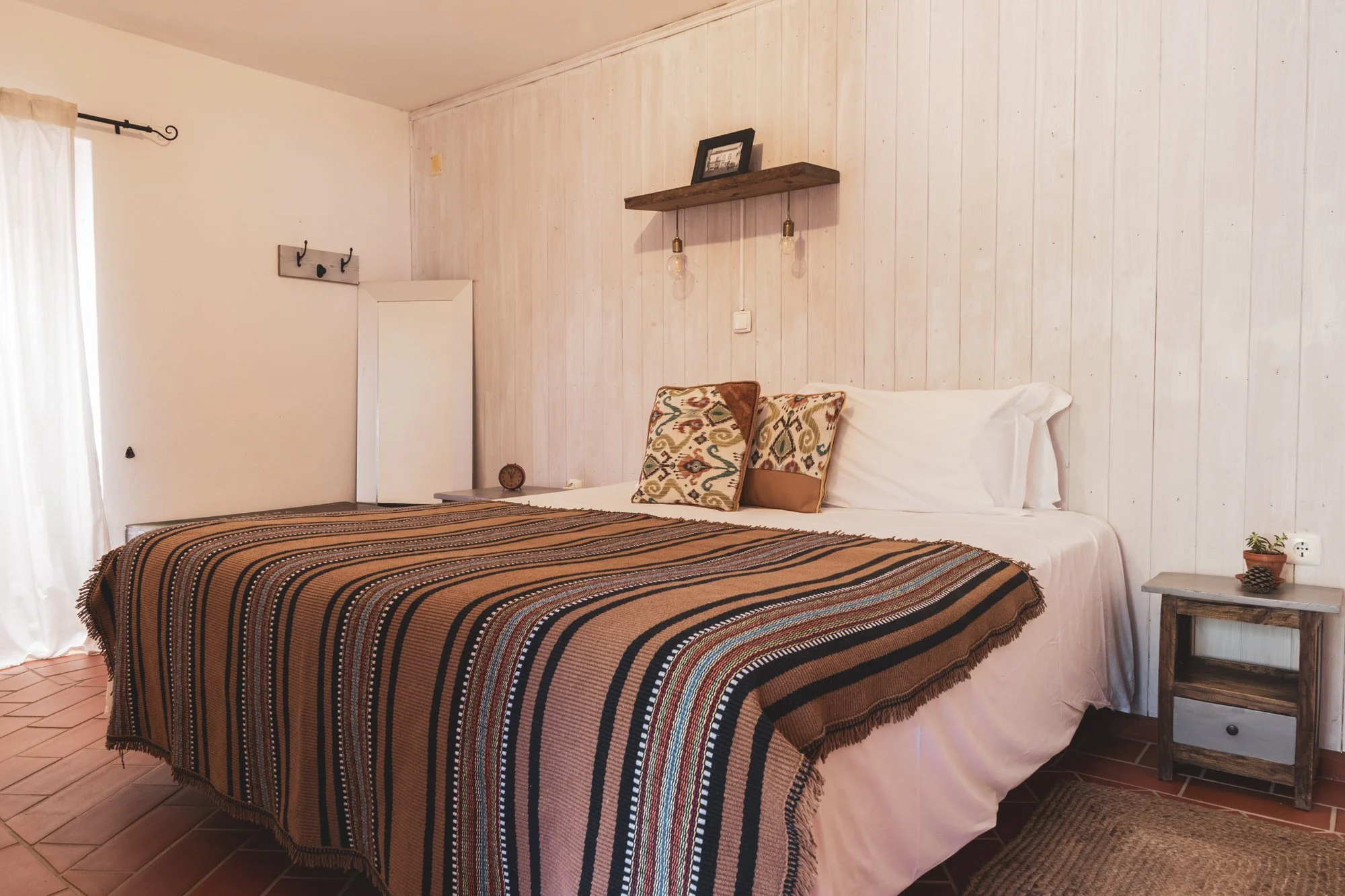 A cozy bedroom with a bed covered in a striped, multicolored blanket. Two decorative pillows with colorful geometric patterns rest against the white headboard. To the right of the bed is a small wooden nightstand with a drawer, on which a small potted plant and a pine cone are placed. The wall behind the bed is paneled in light wood, with a wooden shelf holding a picture frame and two hanging light bulbs. At the left side of the room, a window with sheer curtains allows natural light to illuminate the space.