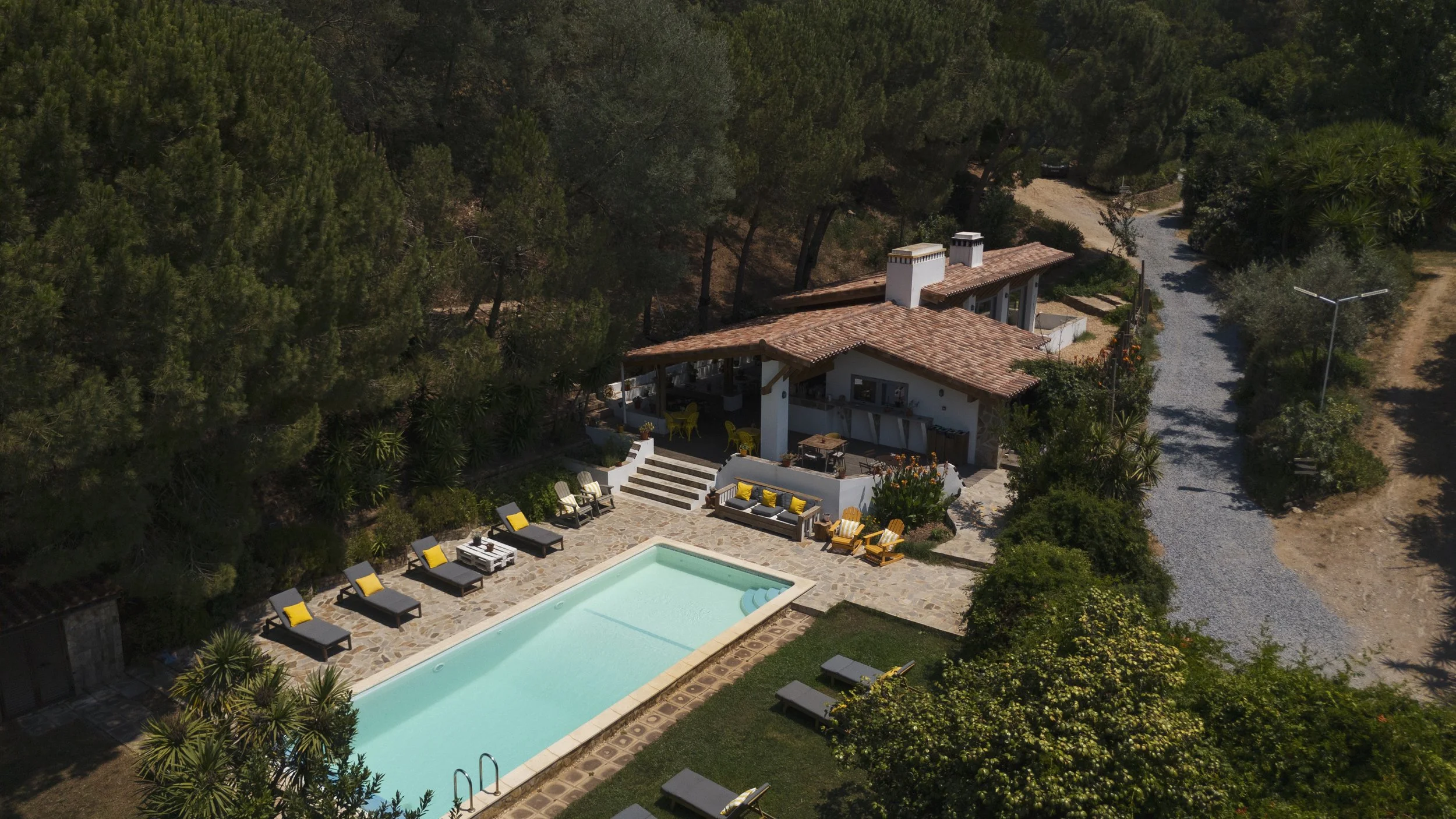 Aerial view of Quinta Camarena Alentejo, Portugal,  with a swimming pool, outdoor lounge area with yellow cushions, surrounded by lush green trees and a rustic house with a tiled roof.