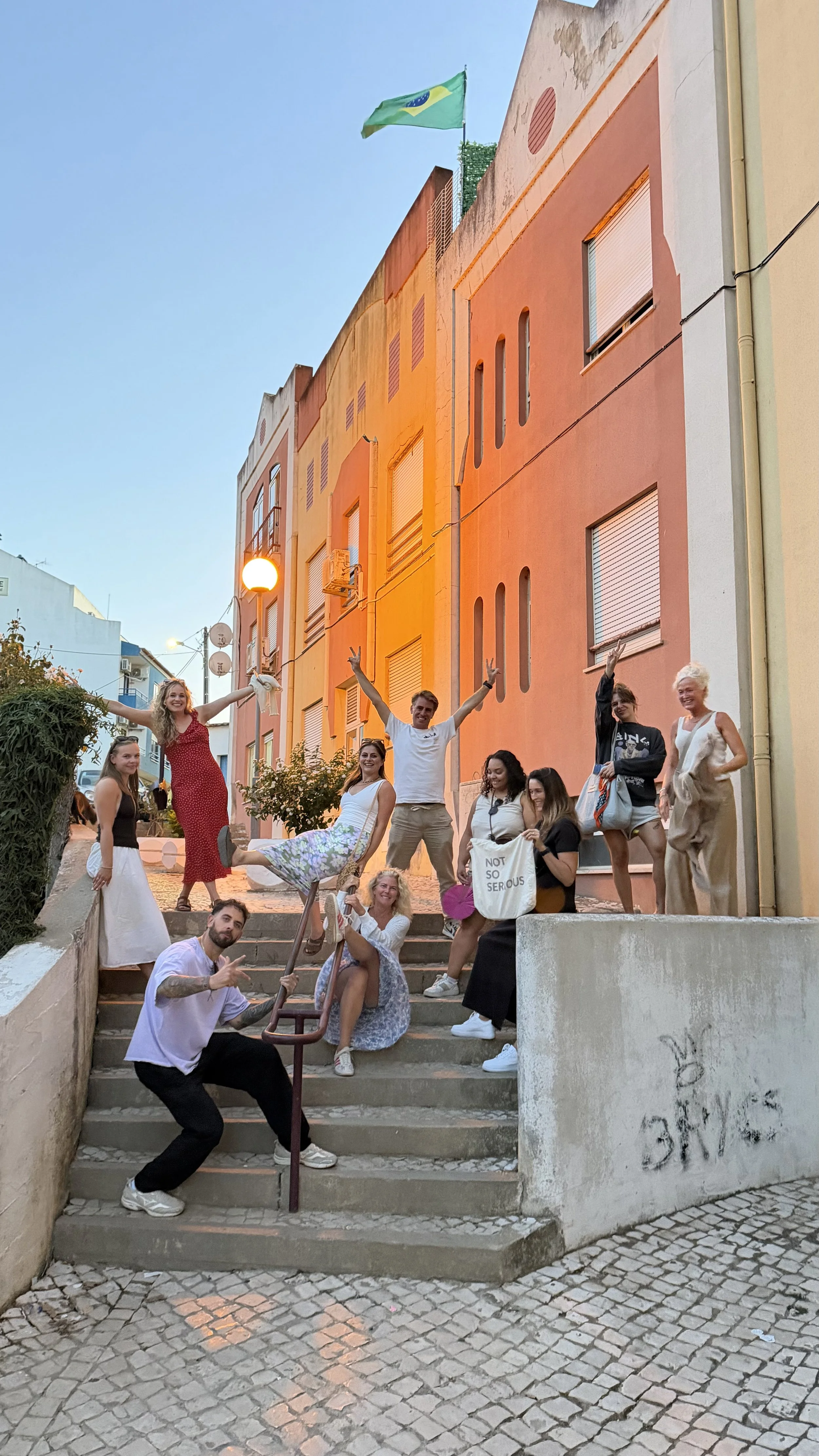 Group of people posing on stairs in an urban street at sunset, with colorful buildings and a Brazilian flag waving in the background.
